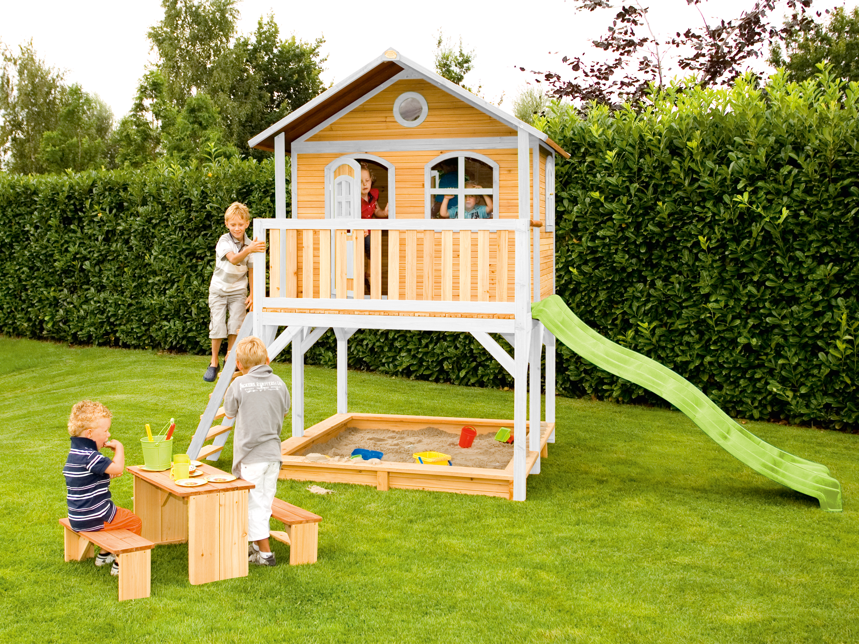 Kinder spielen auf einem Holzspielplatz mit Rutsche, Sandkasten und Spielhaus.