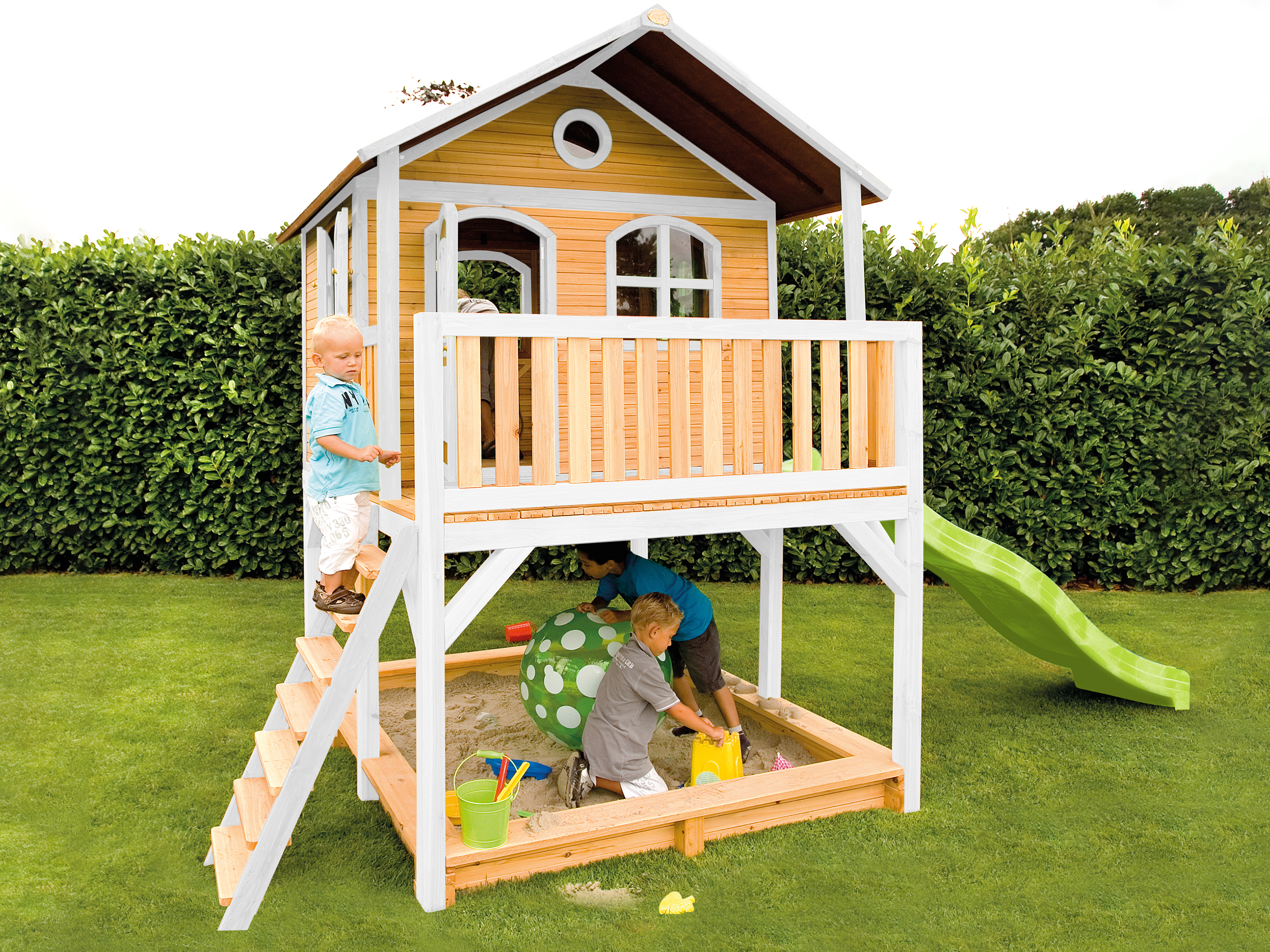 Kinder spielen in einem Sandkasten unter einem Holzspielhaus mit Rutsche. Grünes Gras umgibt es.