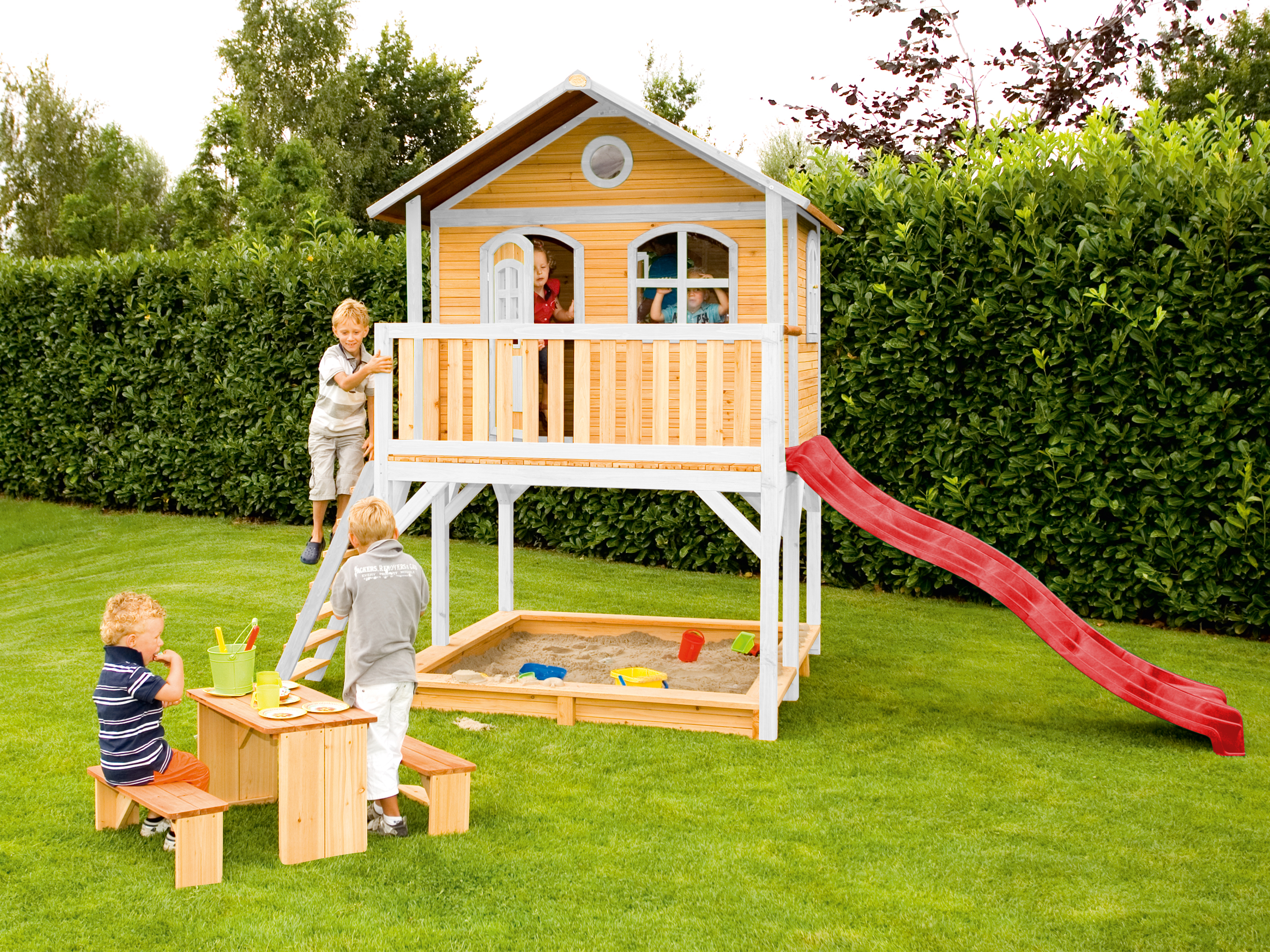 Kinder spielen in einem Holzspielhaus mit Rutsche und Sandkasten in einem grünen Garten.
