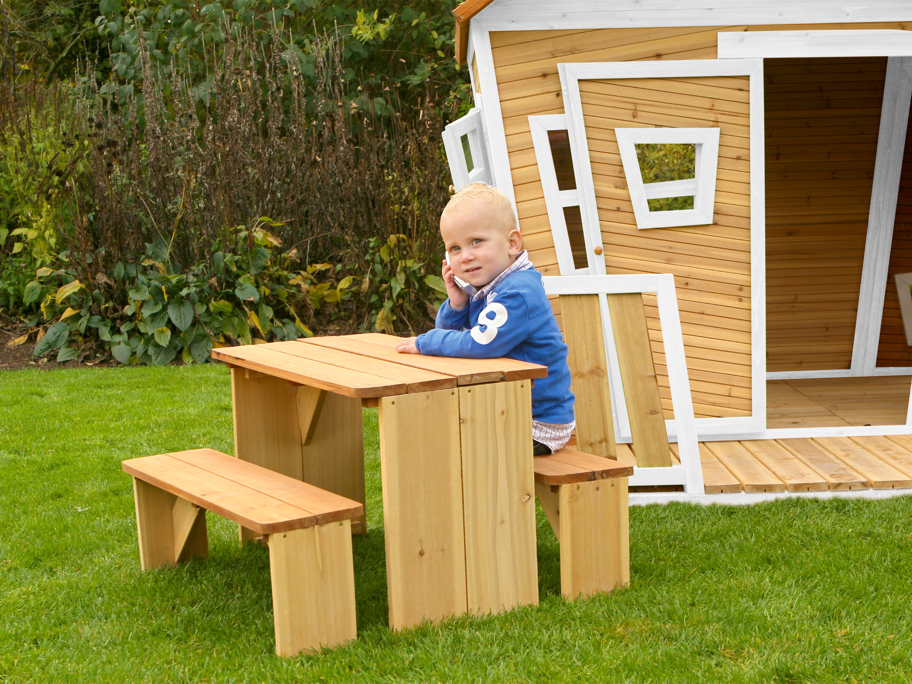 Ein kleiner Junge sitzt an einem kleinen Picknicktisch aus Holz vor einem Spielhaus.