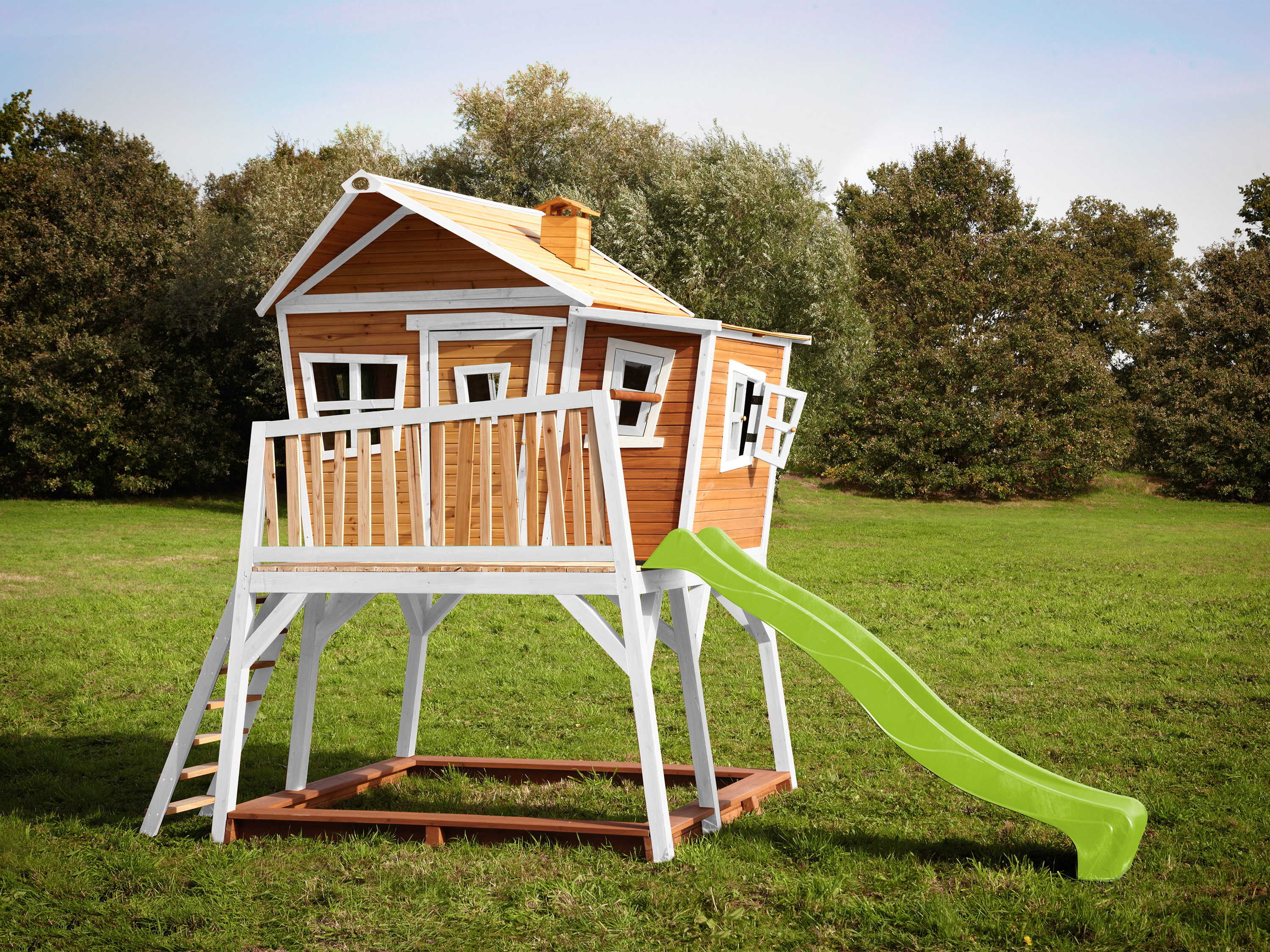 Holzbaumhaus mit grüner Rutsche und weißer Leiter auf einer Wiese, unter blauem Himmel.