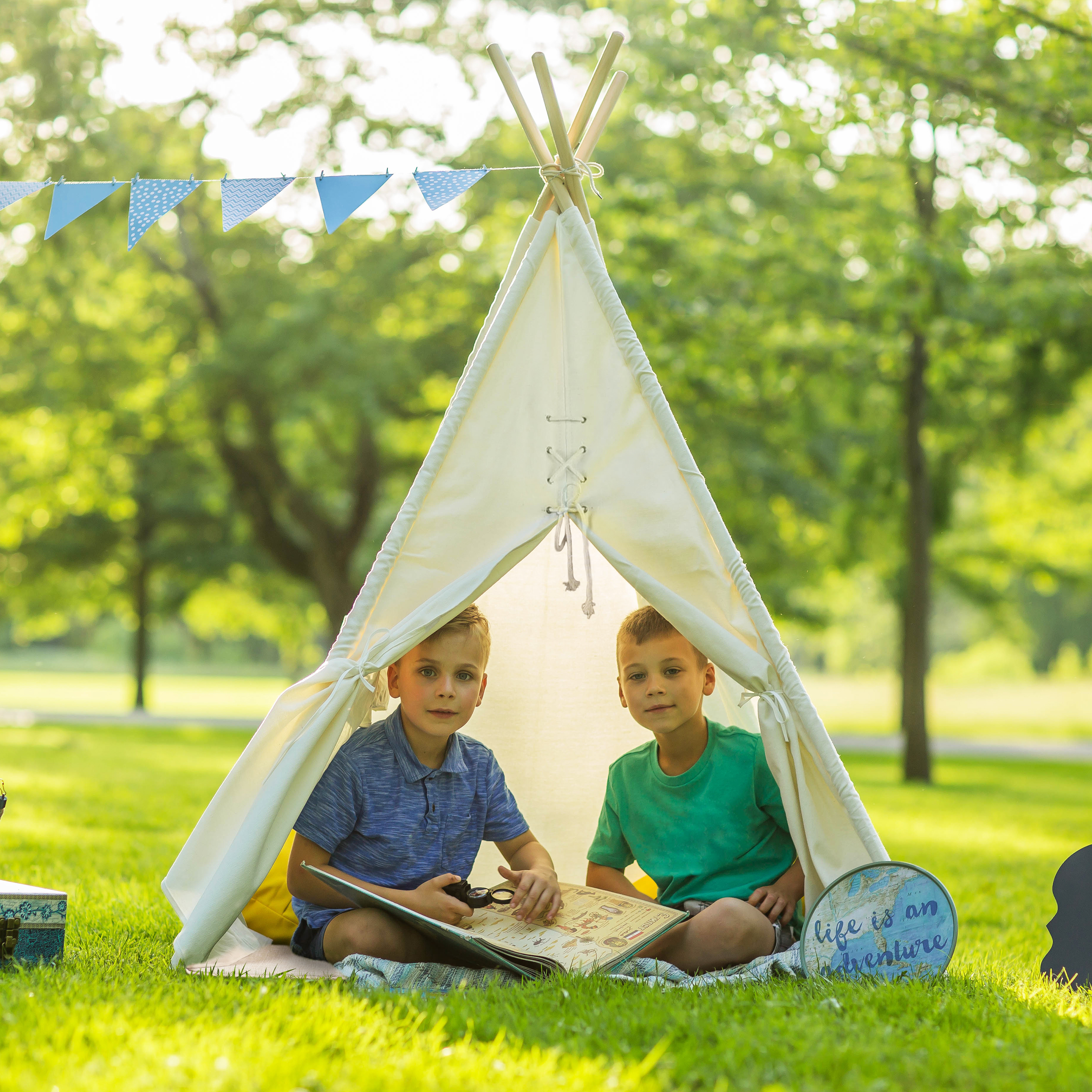 Zwei Jungen lesen in einem Zelt ein Buch im Park, mit Banner und Schild in der Nähe. Grünes Gras.