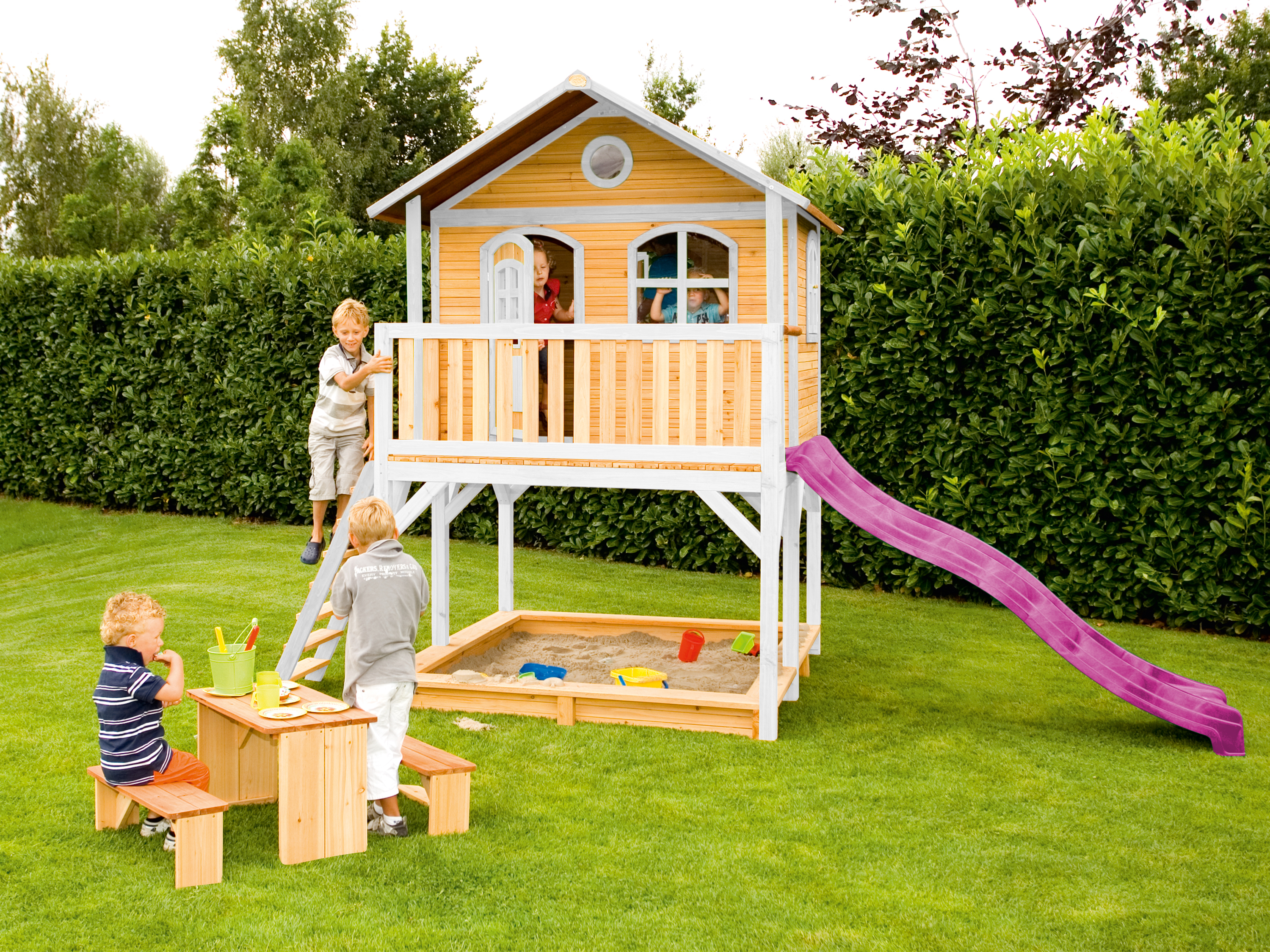 Kinder spielen auf einem hölzernen Spielhaus mit Rutsche in einem grünen Garten.