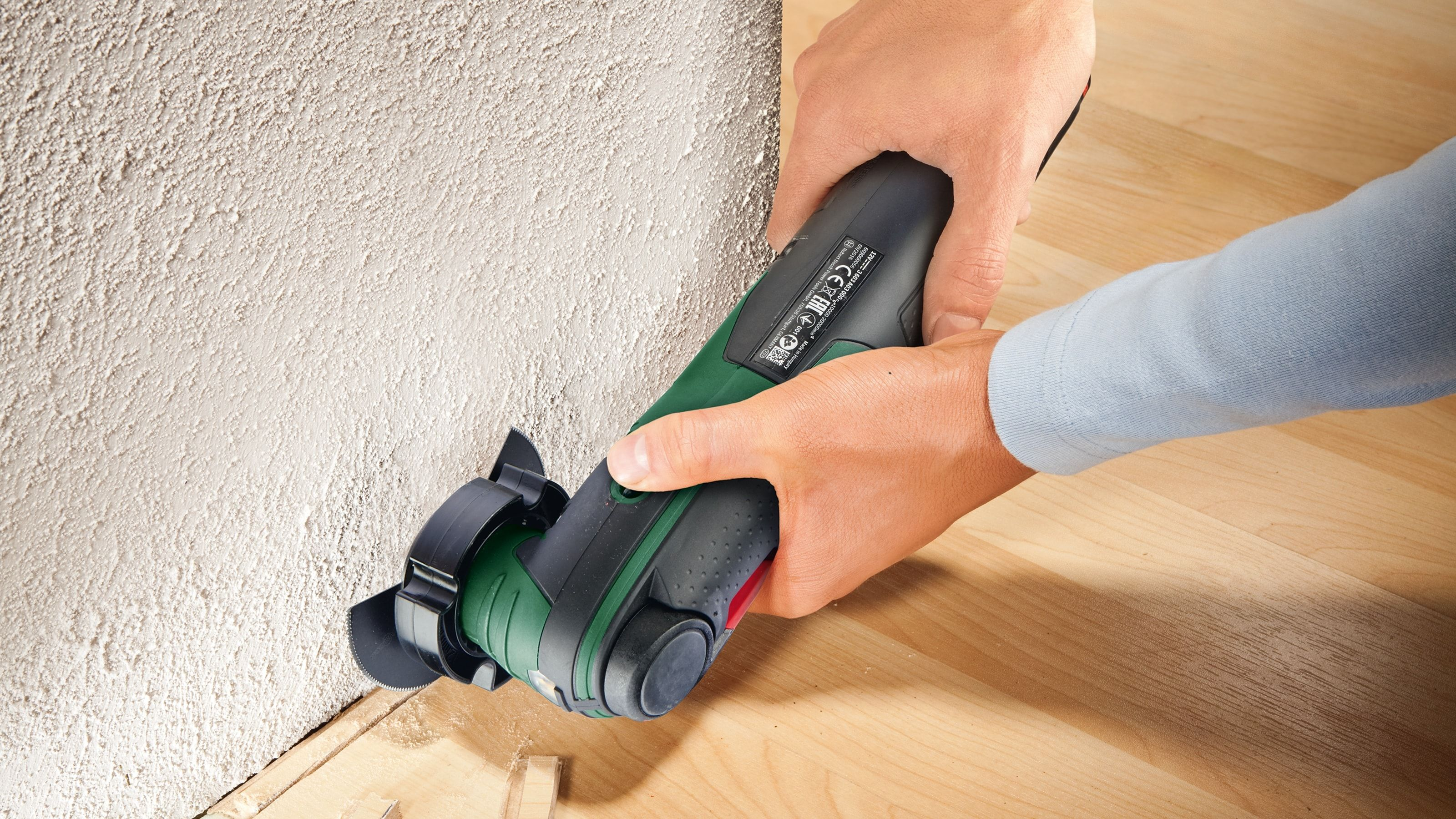 Hände halten ein grün-schwarzes Multitool, das eine Wandkante in der Nähe eines Holzbodens schneidet.
