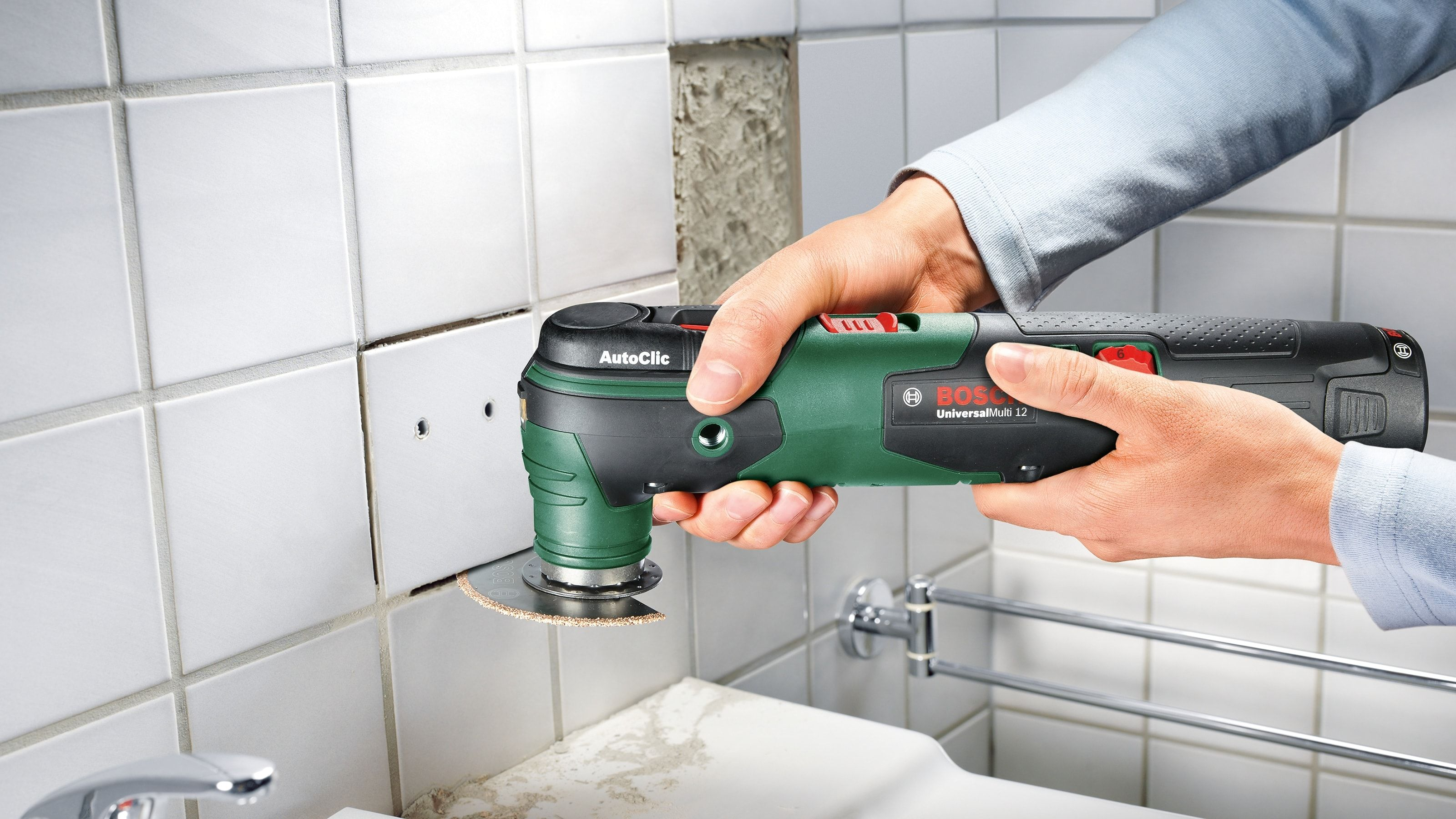 Hand, die ein grün-schwarzes Multitool verwendet, um Fliesen in der Nähe eines Waschbeckens und einer weißen Wand zu schneiden.