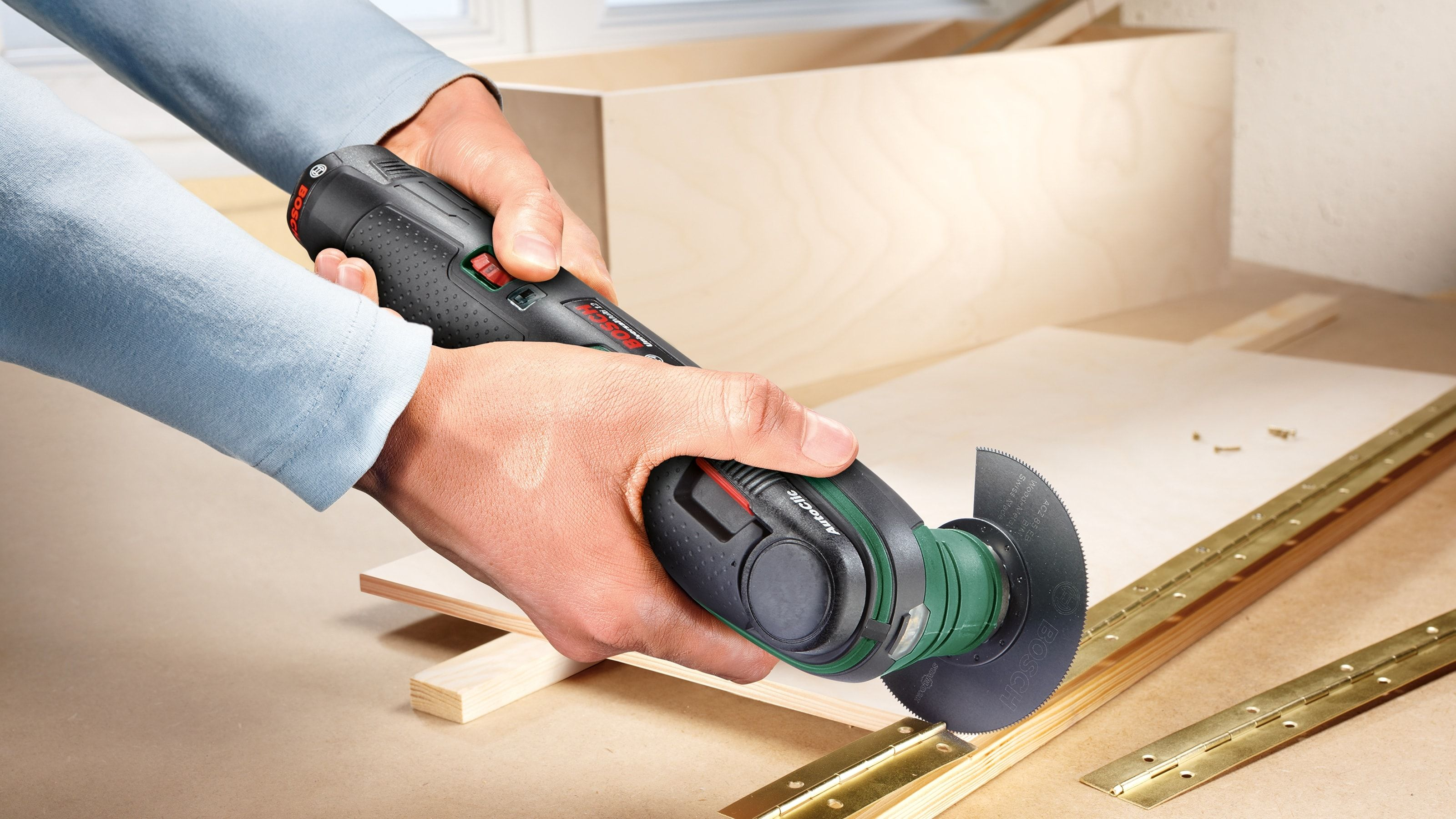 Hände halten ein grün-schwarzes Multitool, das Holz in der Nähe einer Sperrholzplatte schneidet.
