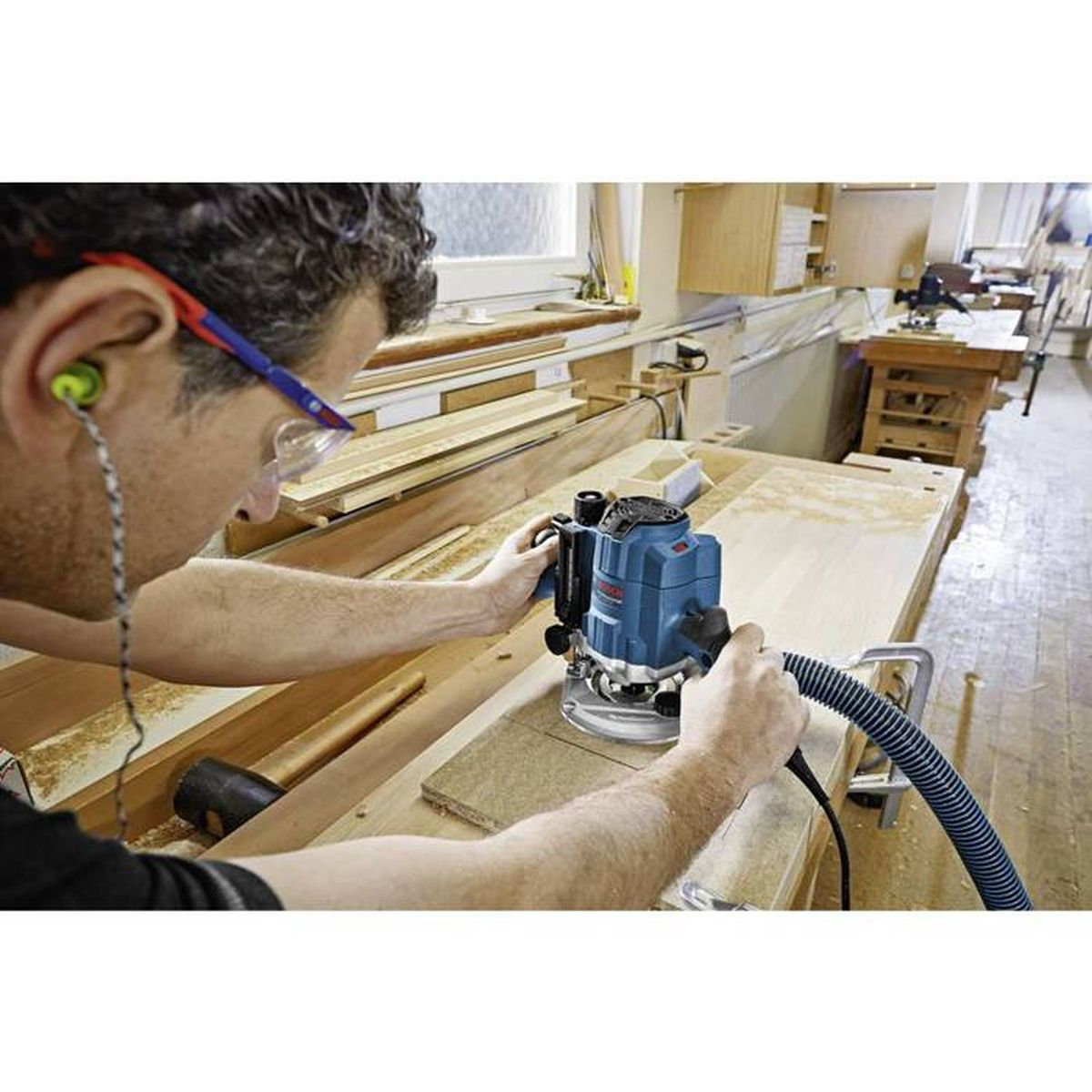 Mężczyzna używa frezarki Bosch na drewnianej powierzchni. Nosi okulary ochronne i ochronniki słuchu.