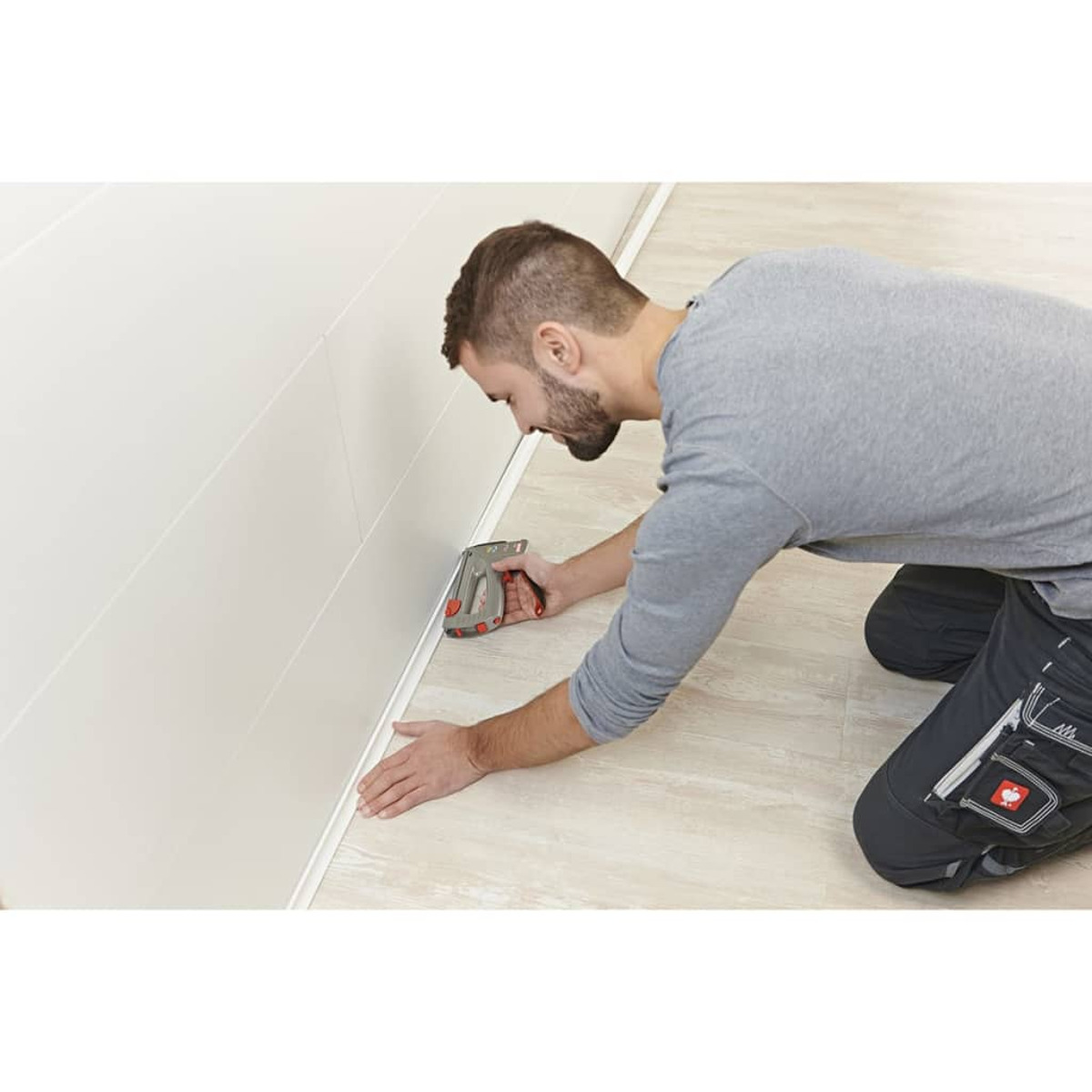 Un uomo è in ginocchio, usa una pinzatrice per fissare un elemento di finitura a un muro bianco e un pavimento chiaro.