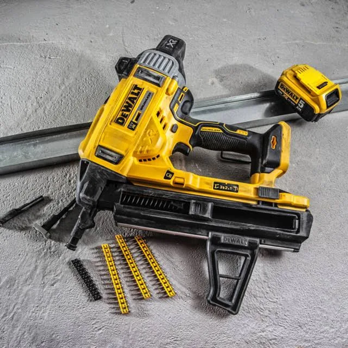 Una sparachiodi DeWalt gialla e un pacco batteria su una superficie grigia con chiodi e materiali da costruzione.