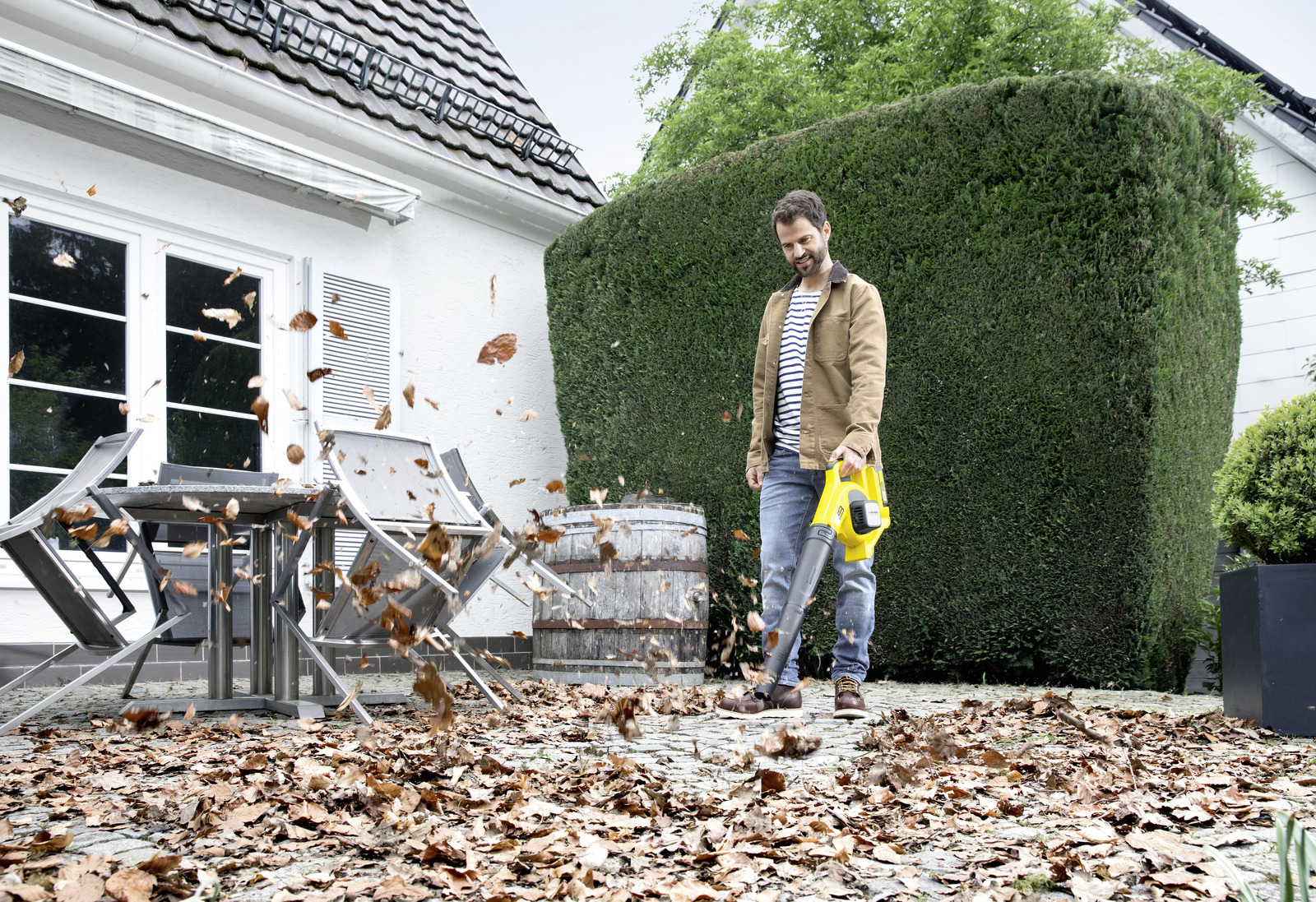 Un homme utilisant un souffleur de feuilles sur une terrasse couverte de feuilles, avec une maison et une haie en arrière-plan.