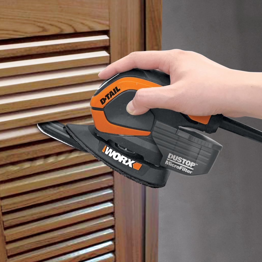 Mano che leviga una porta di legno con una levigatrice Worx D-Tail arancione e nera. Primo piano.