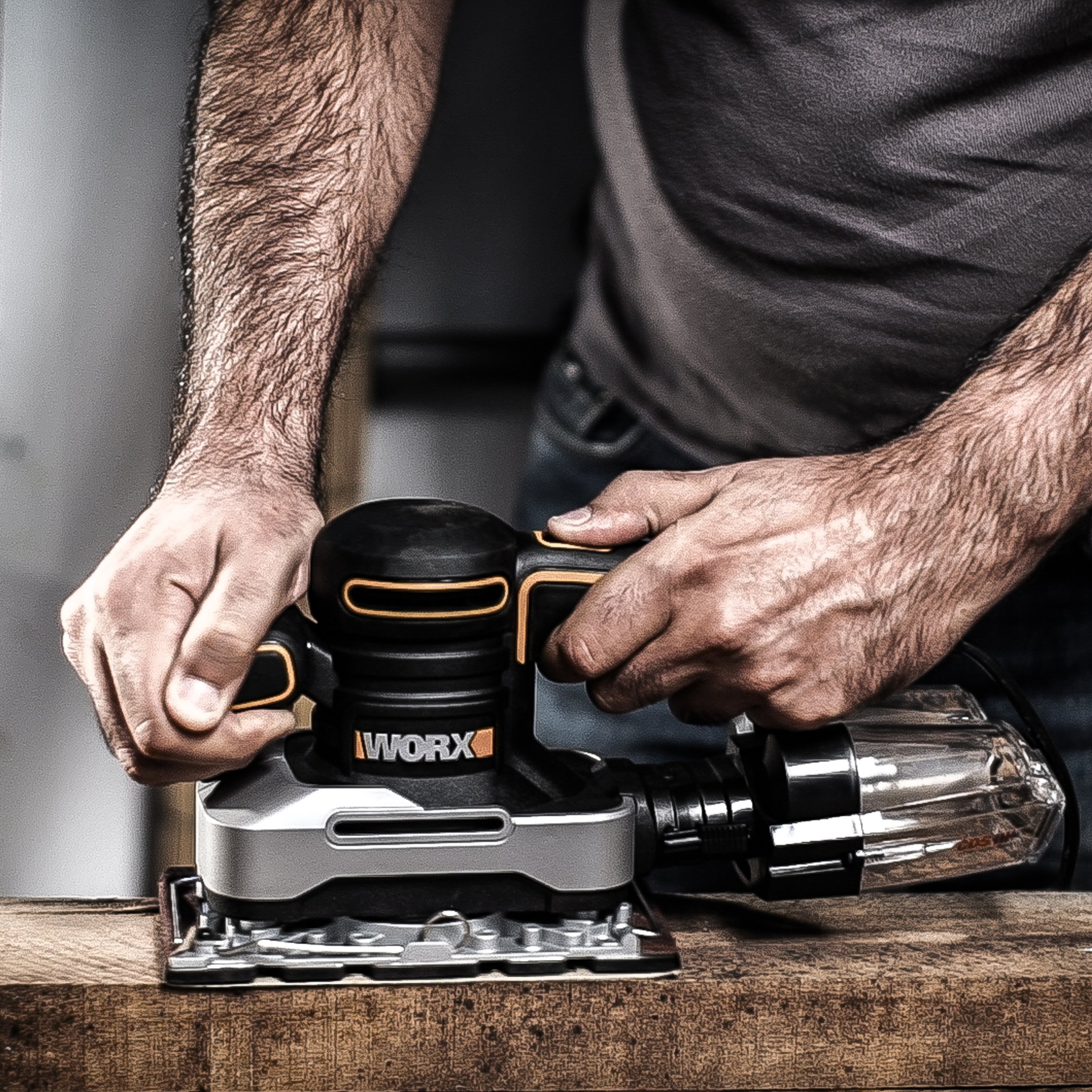 Mani che tengono una levigatrice WORX nera e argento su una superficie di legno. Raccoglitore polvere.