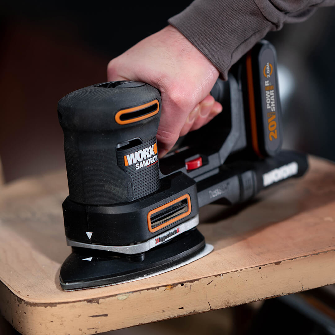 Ręka osoby trzymająca szlifierkę na jasnobrązowej drewnianej powierzchni. Marka to WORX.