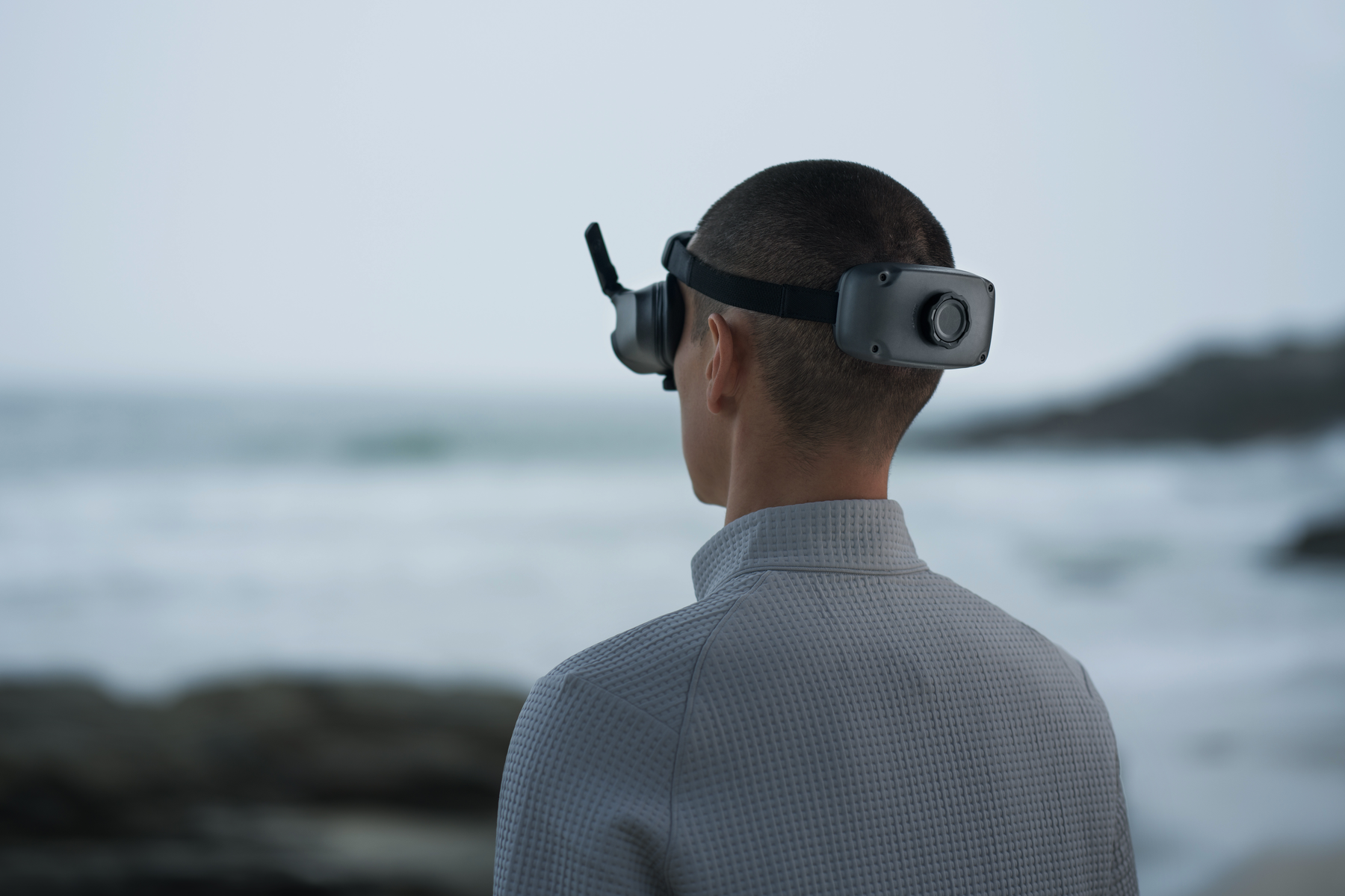 Un uomo con un visore VR, di spalle alla fotocamera, guarda il mare. Cielo nuvoloso.