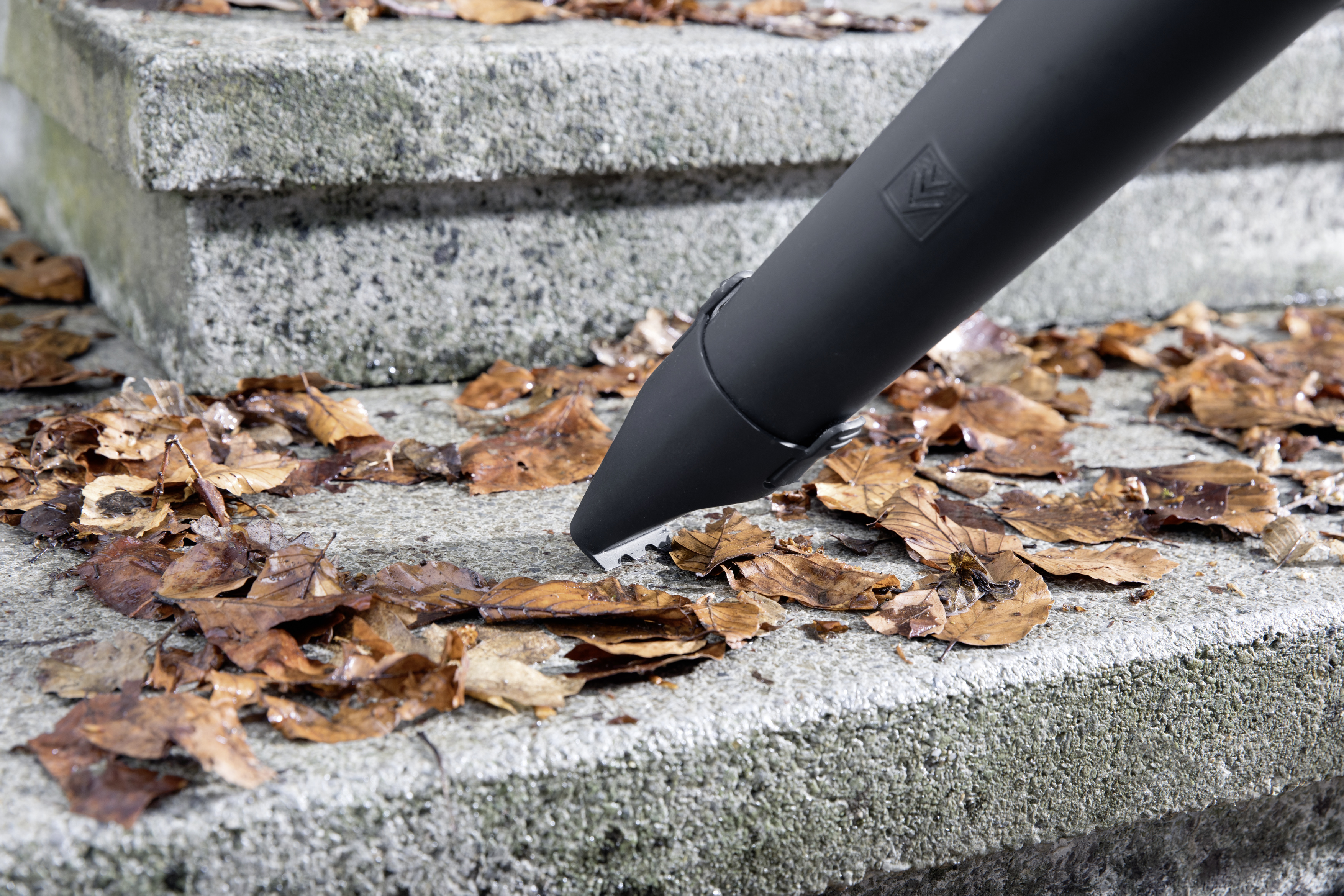 Dysza dmuchawy do liści zdmuchująca liście ze stopni kamiennych. Dominują kolory brązowy i szary.