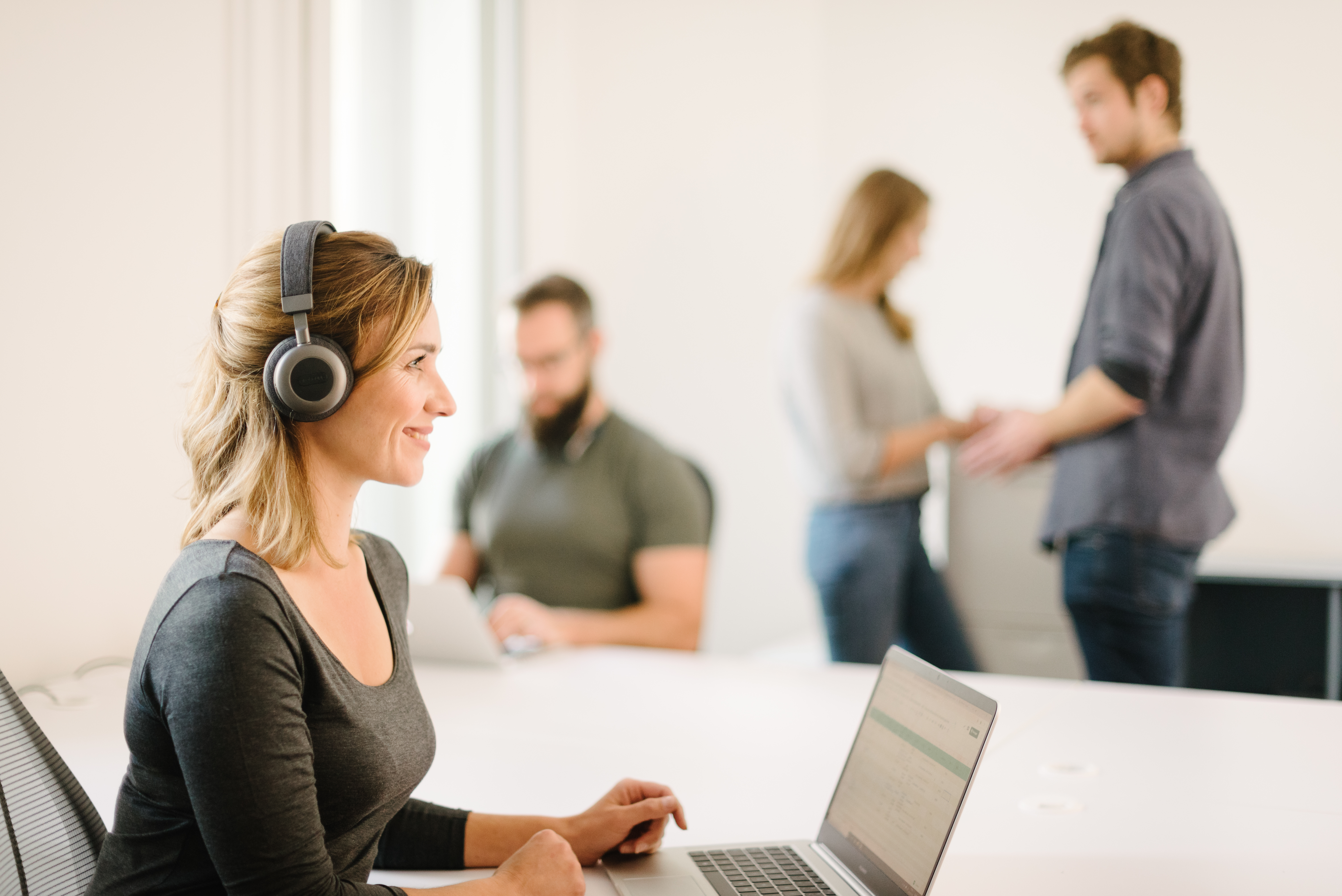 Woman wearing headphones and using a laptop in an office. People in the background.