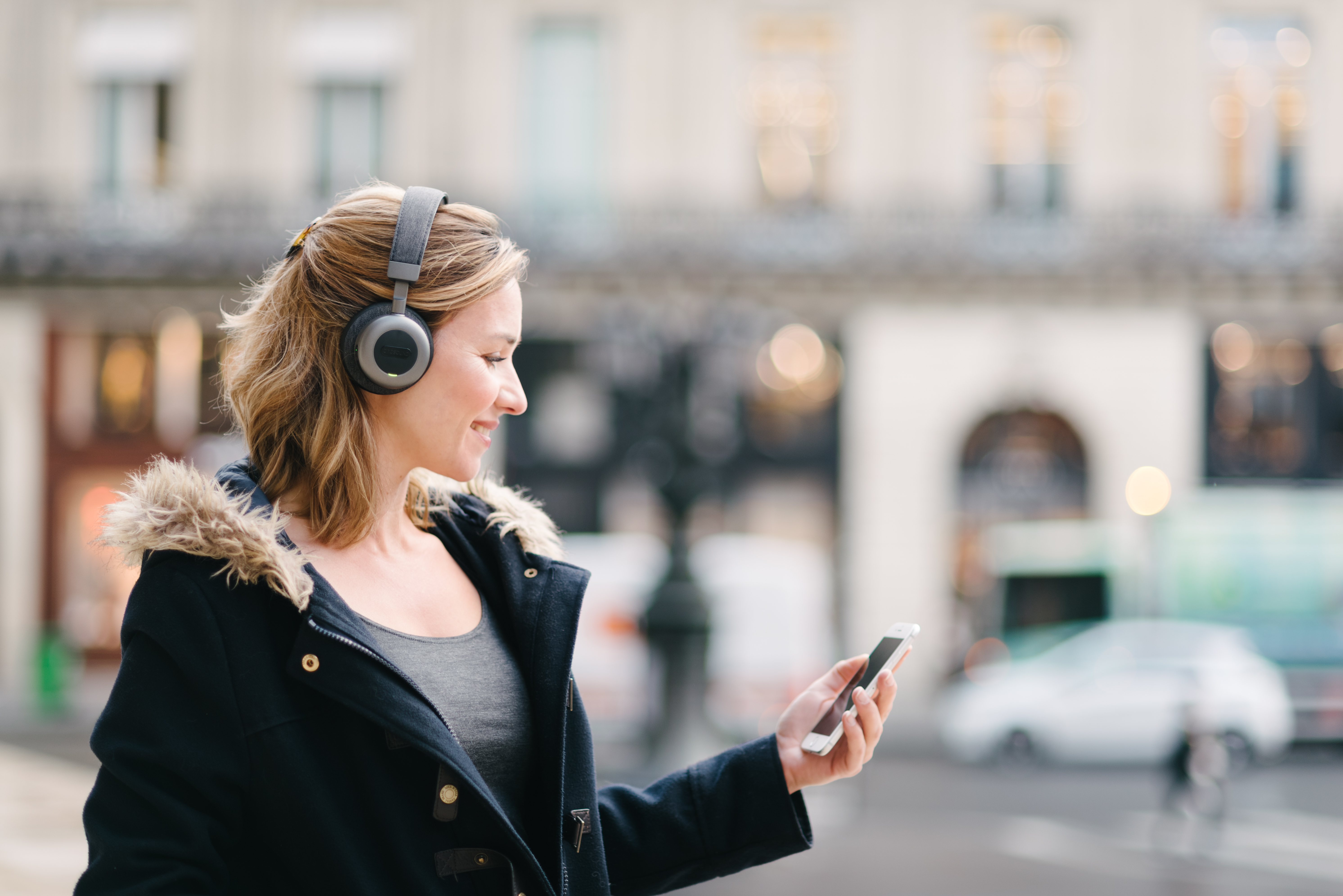 Woman wearing headphones and looking at her phone on a city street. Buildings in background.