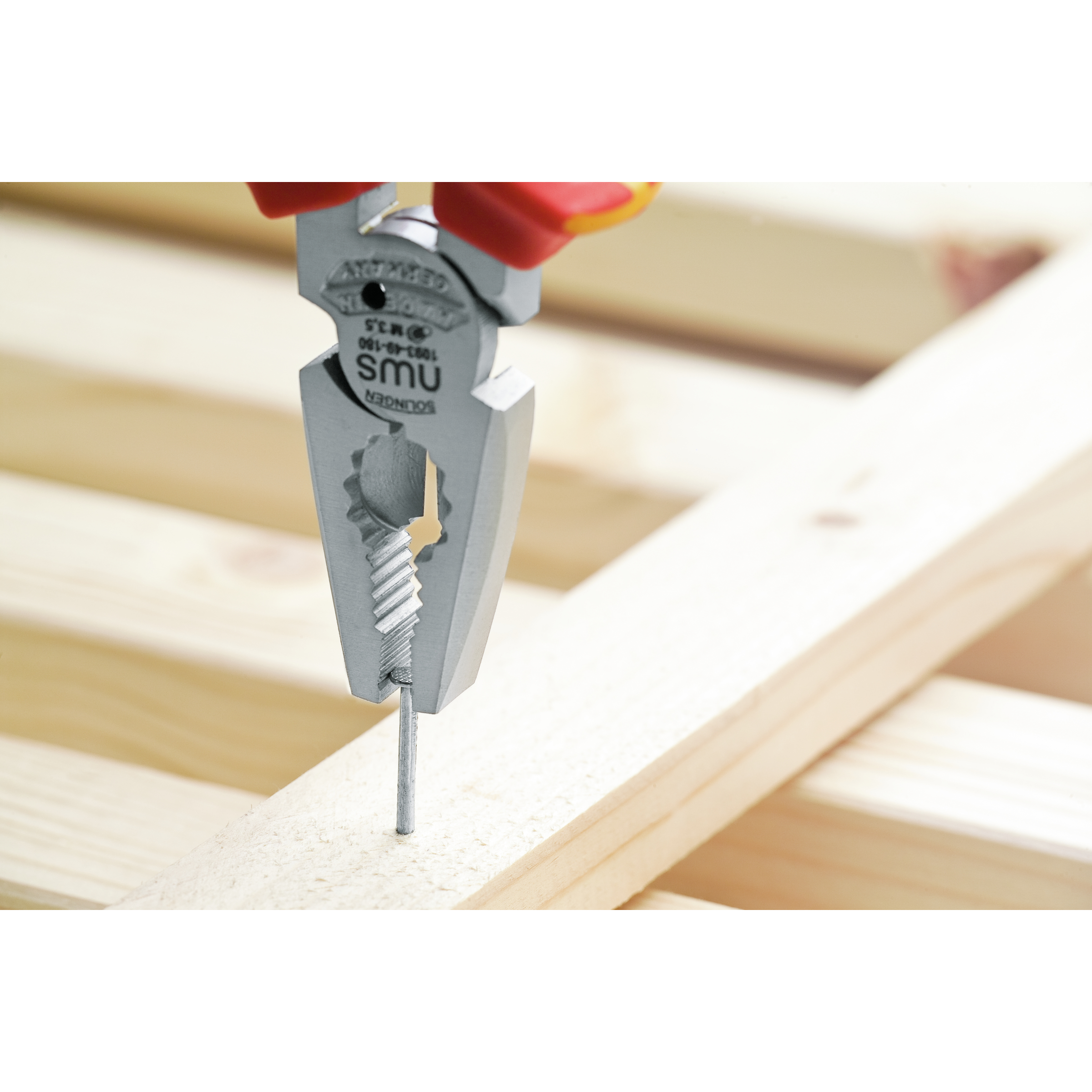 Szczypce trzymające gwóźdź na drewnie. Czerwono-żółte uchwyty, srebrne szczęki. Drewniane tło.