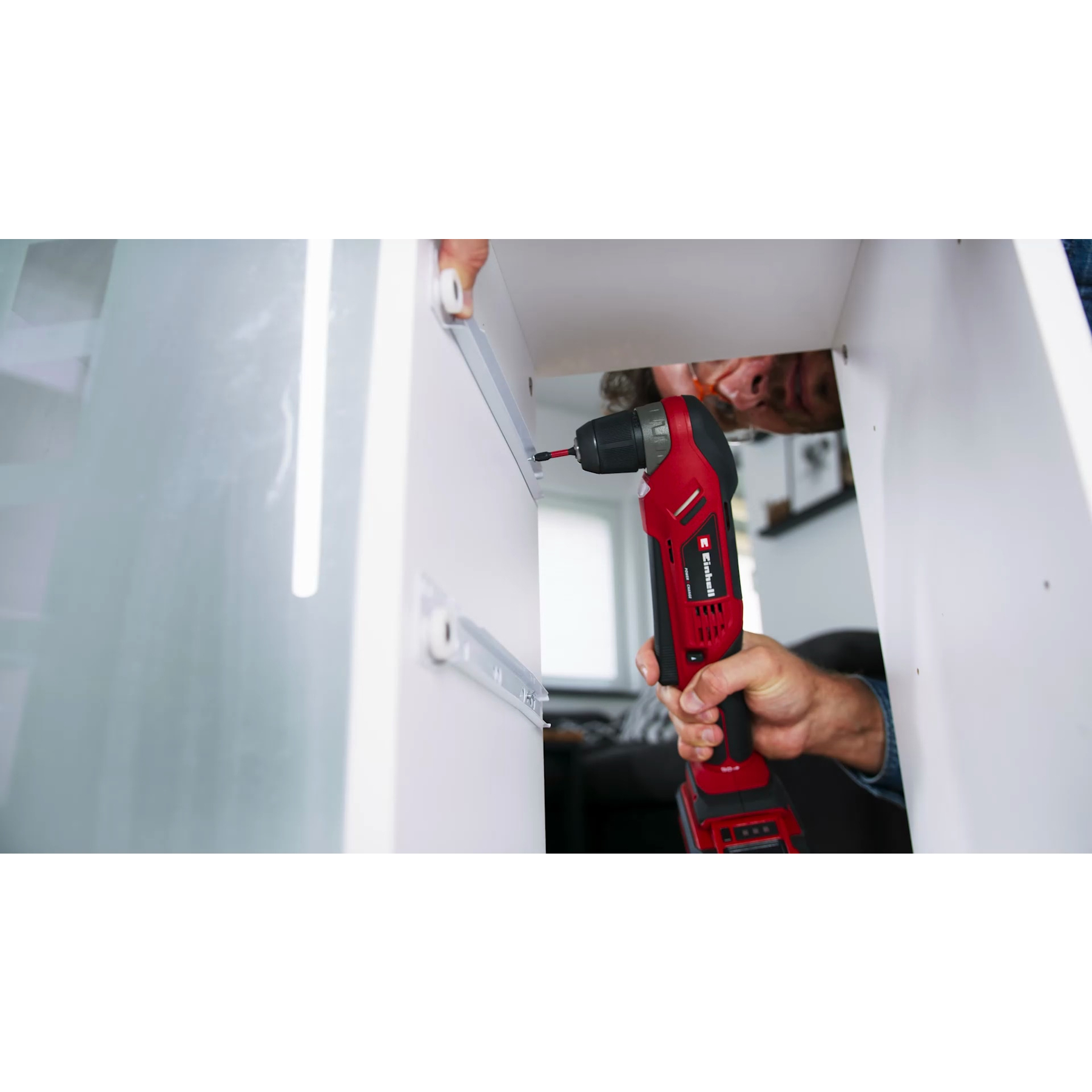 Mężczyzna używa czerwono-czarnej wiertarko-wkrętarki do montażu białej szafki.