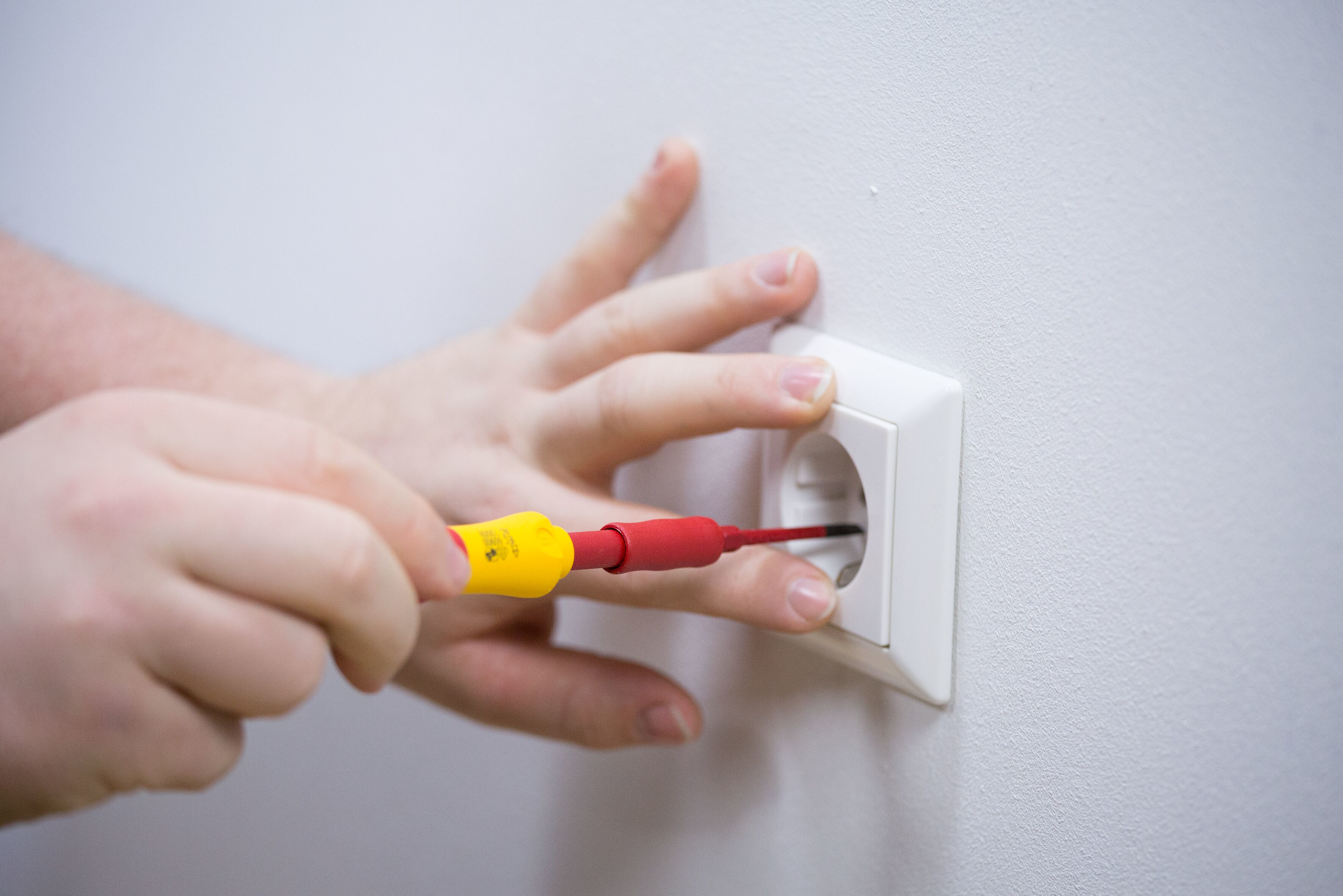 Osoba używająca czerwono-żółtego śrubokrętu do gniazdka elektrycznego.