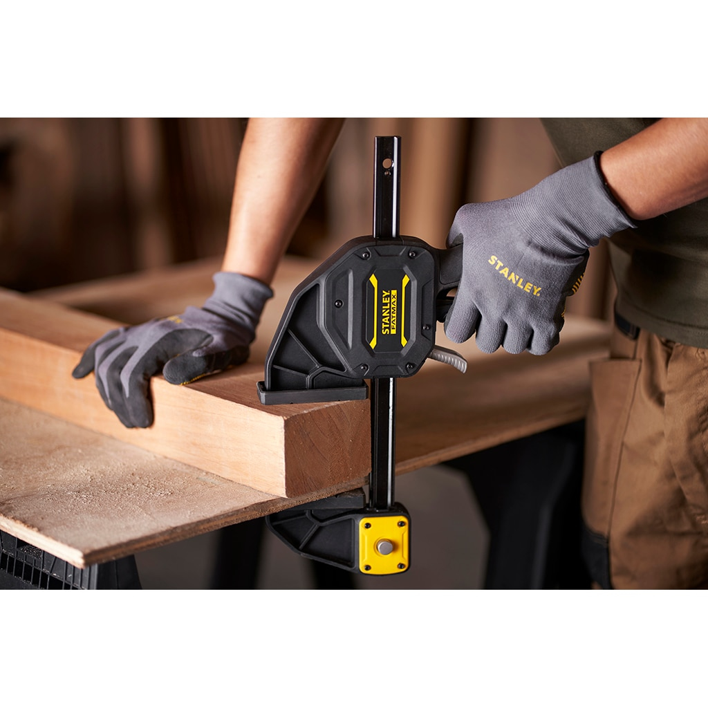 Una persona utiliza una abrazadera Stanley negra y amarilla para asegurar una viga de madera. La abrazadera se usa en una superficie de trabajo.
