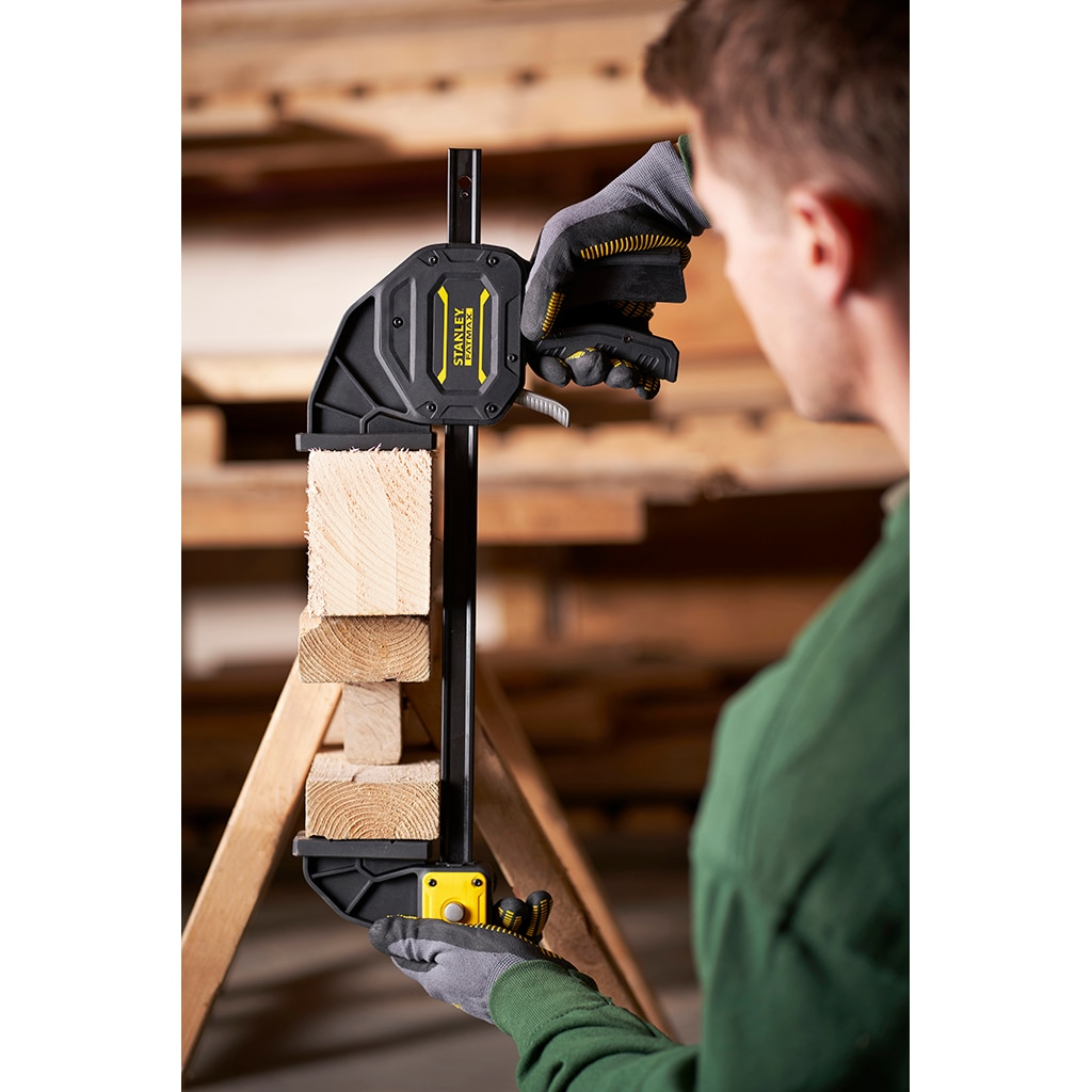 Una persona usa una abrazadera Stanley Fatmax en madera. La abrazadera es negra y amarilla. La madera está en un marco de madera.