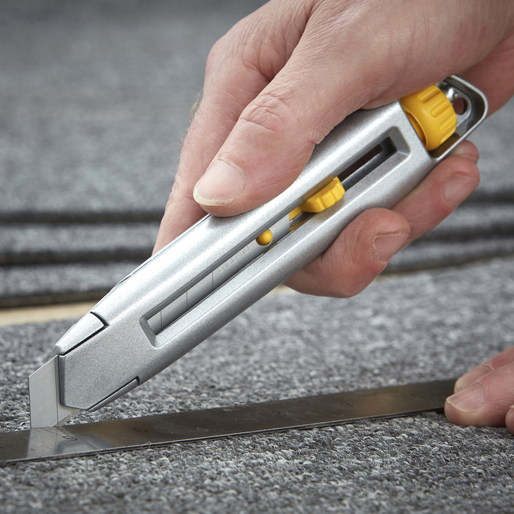 Metal bir cetvelle halı kesmek için gümüş bir maket bıçağı kullanan kişi. Sarı detaylar.