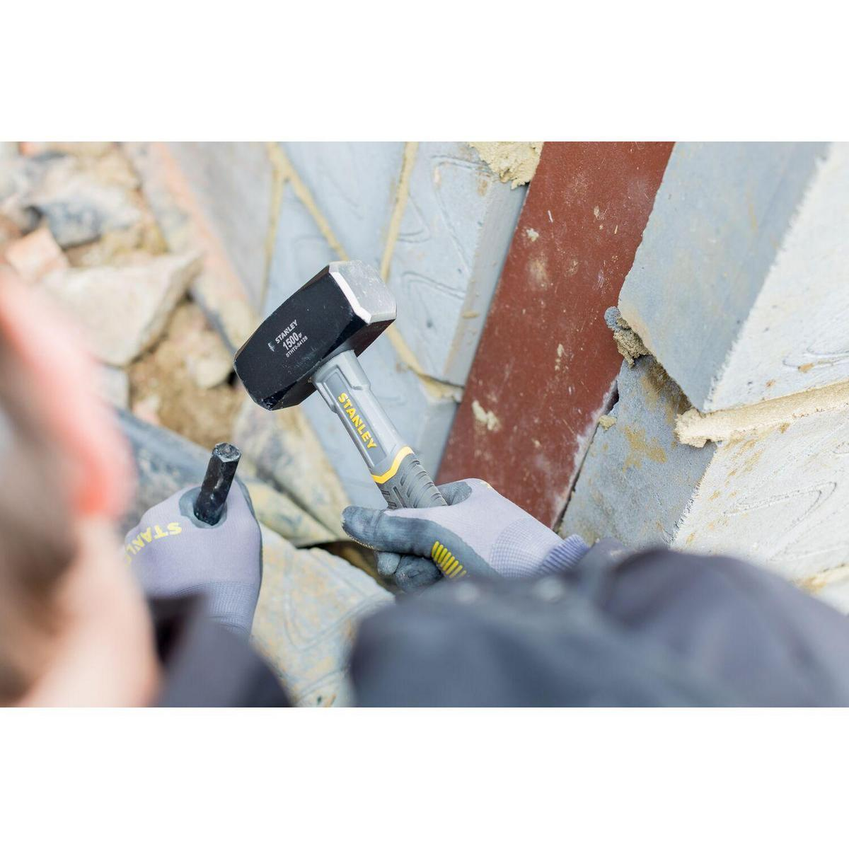 Eine Person benutzt einen Vorschlaghammer und ein Werkzeug, um auf einen Steinblock in der Nähe einer Backsteinmauer zu schlagen.