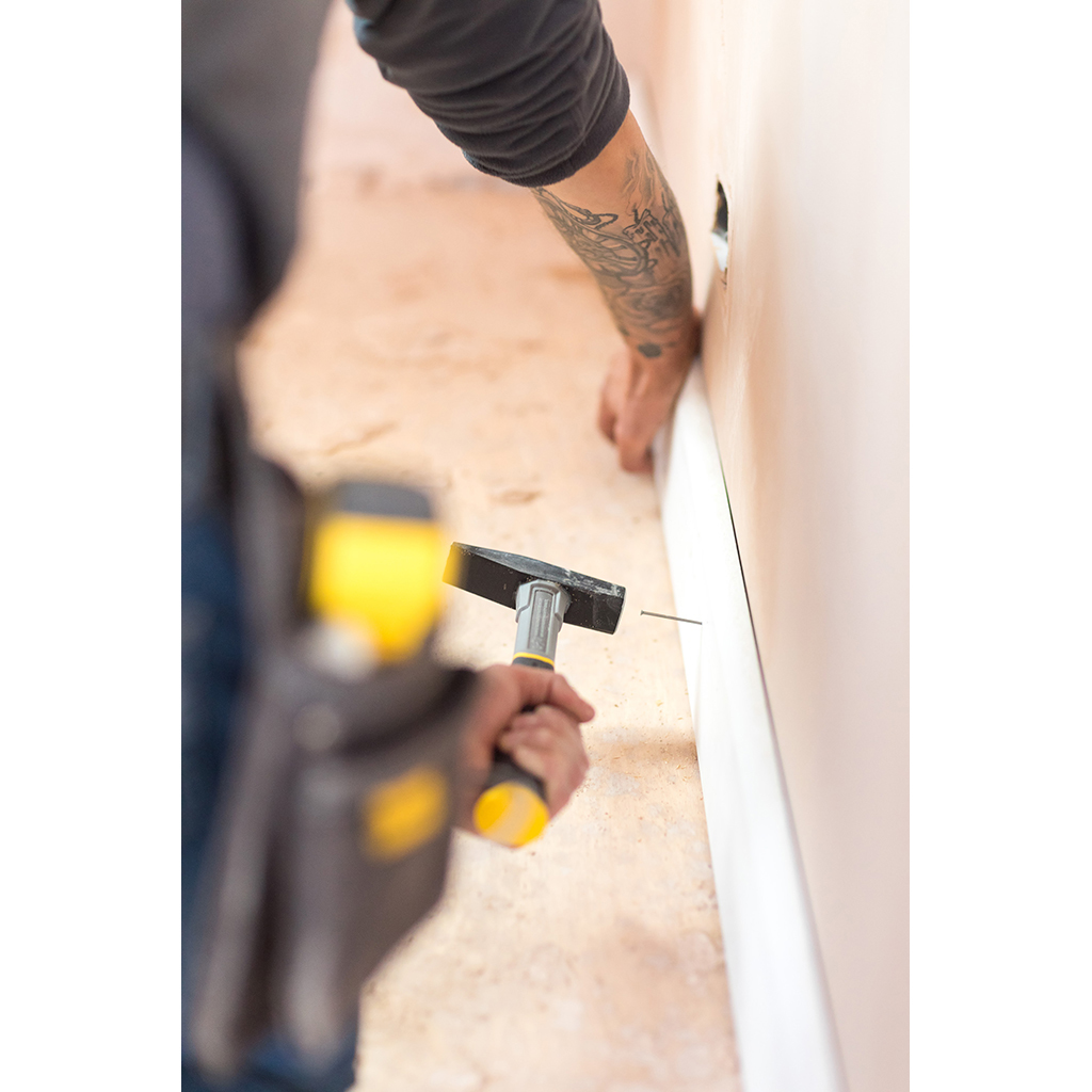 Osoba wbija gwóźdź w białą deskę. Używany jest młotek z żółtą rączką.