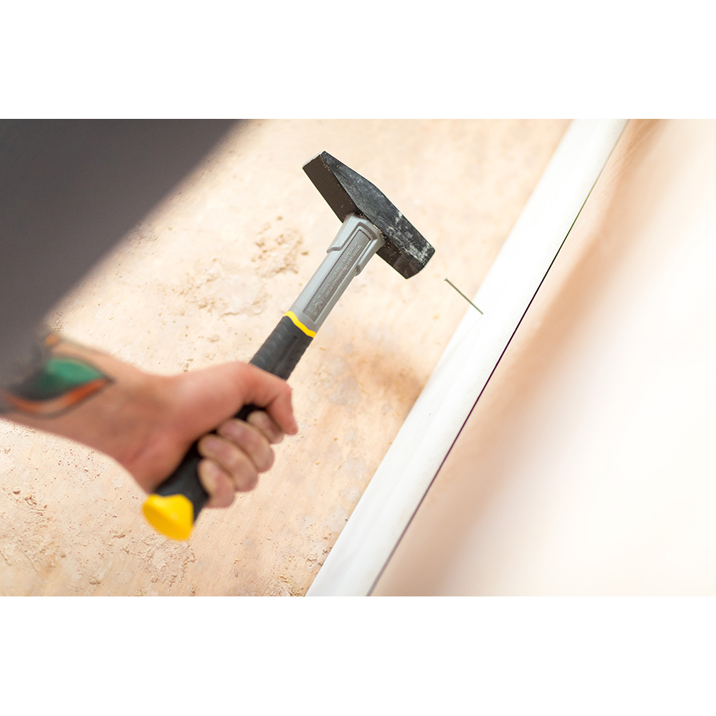 Osoba wbija gwóźdź w białą deskę. Używany jest młotek z żółtą rączką.