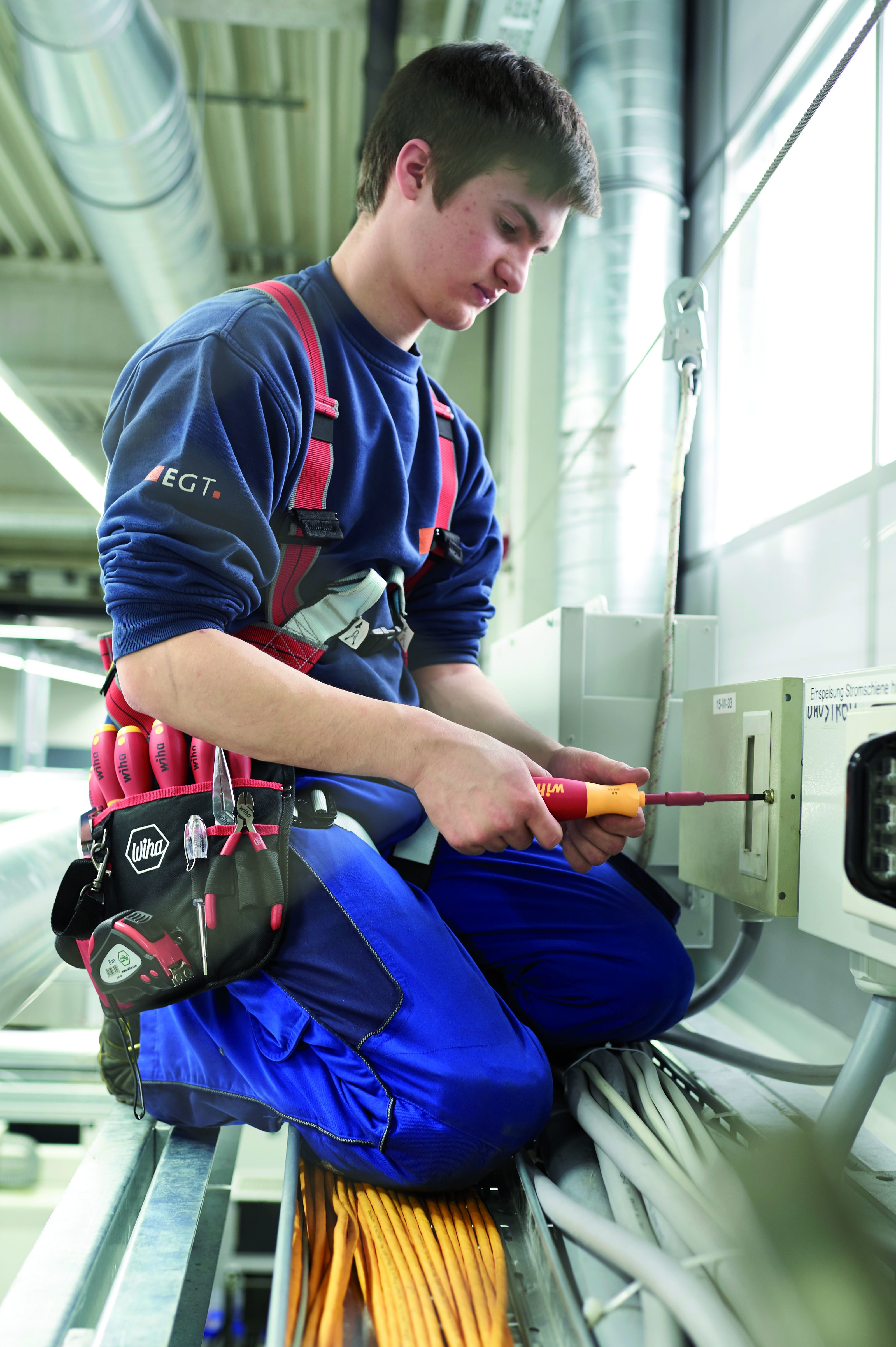Hombre de rodillas trabajando con cables. Usa ropa de trabajo, arneses y un destornillador.