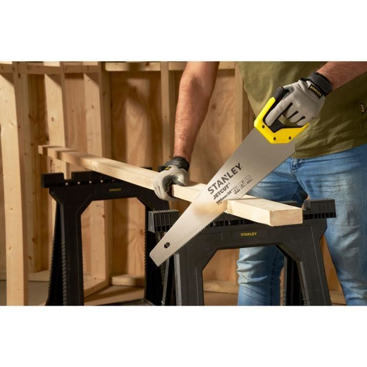 Osoba tnie drewno piłą ręczną. Czarne kozły w tle. Drewniana konstrukcja.