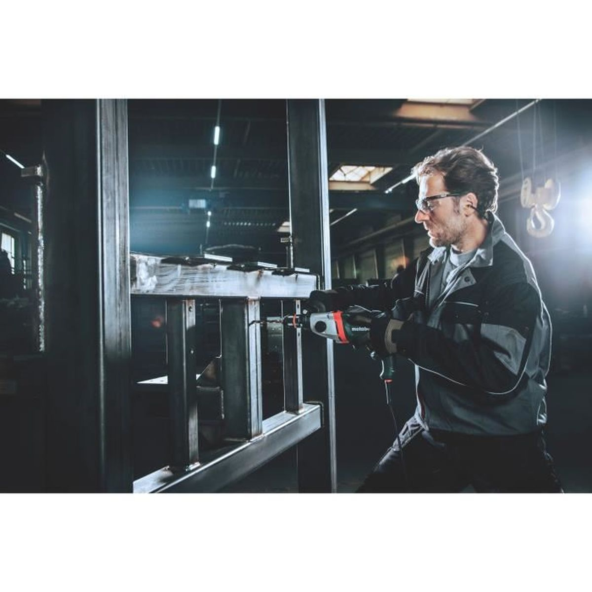 Mężczyzna w okularach używa wiertarki na metalowej konstrukcji. Szara kurtka i okulary.