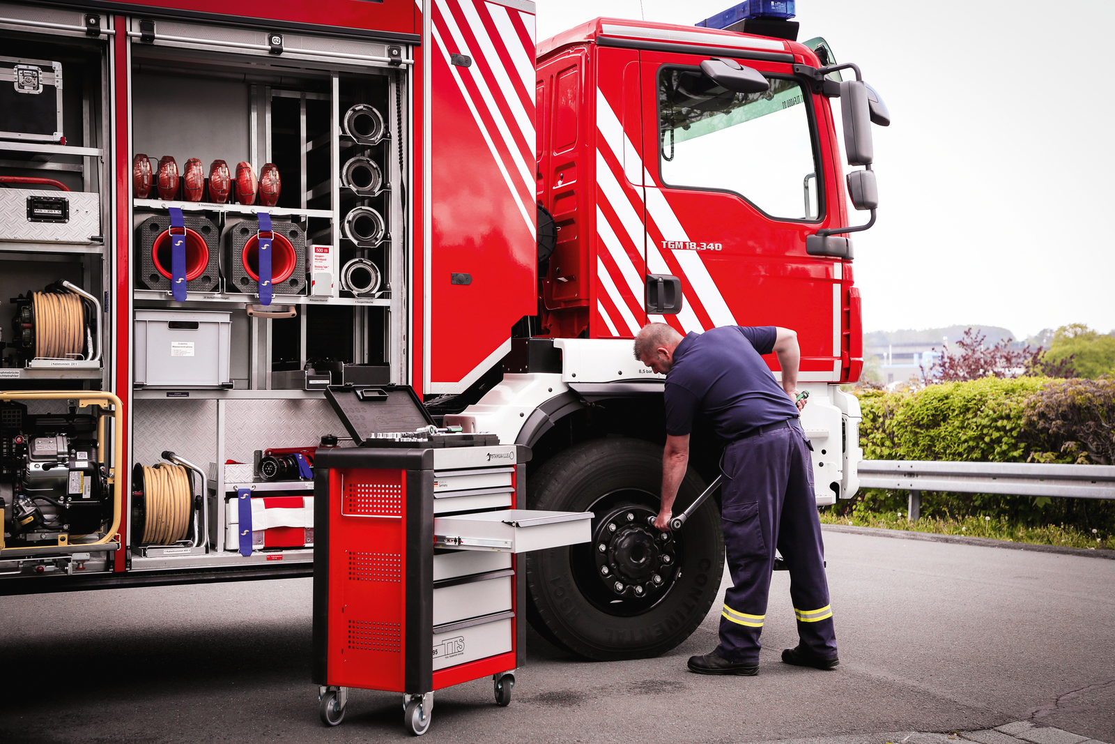 Feuerwehrwagen, rot und weiß, mit einem Mann, der an einem Reifen arbeitet, Werkzeugkasten in der Nähe.