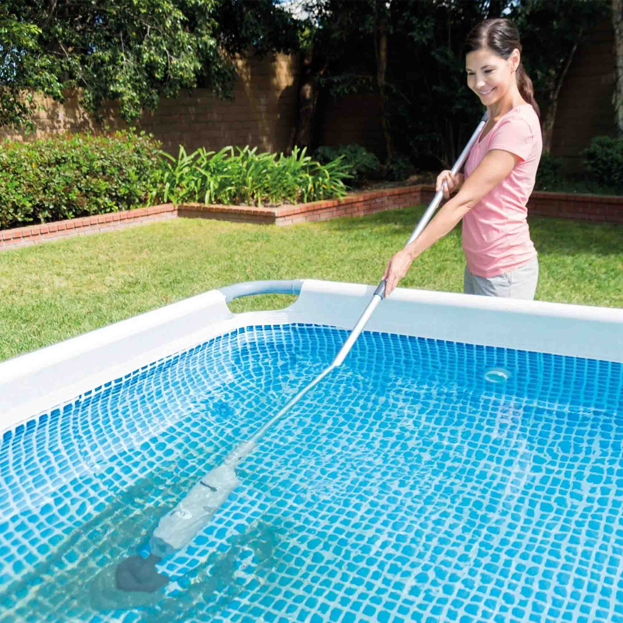 Frau reinigt einen Pool mit einem Staubsauger mit langem Griff. Blauer Pool, grünes Gras, sonniger Tag.