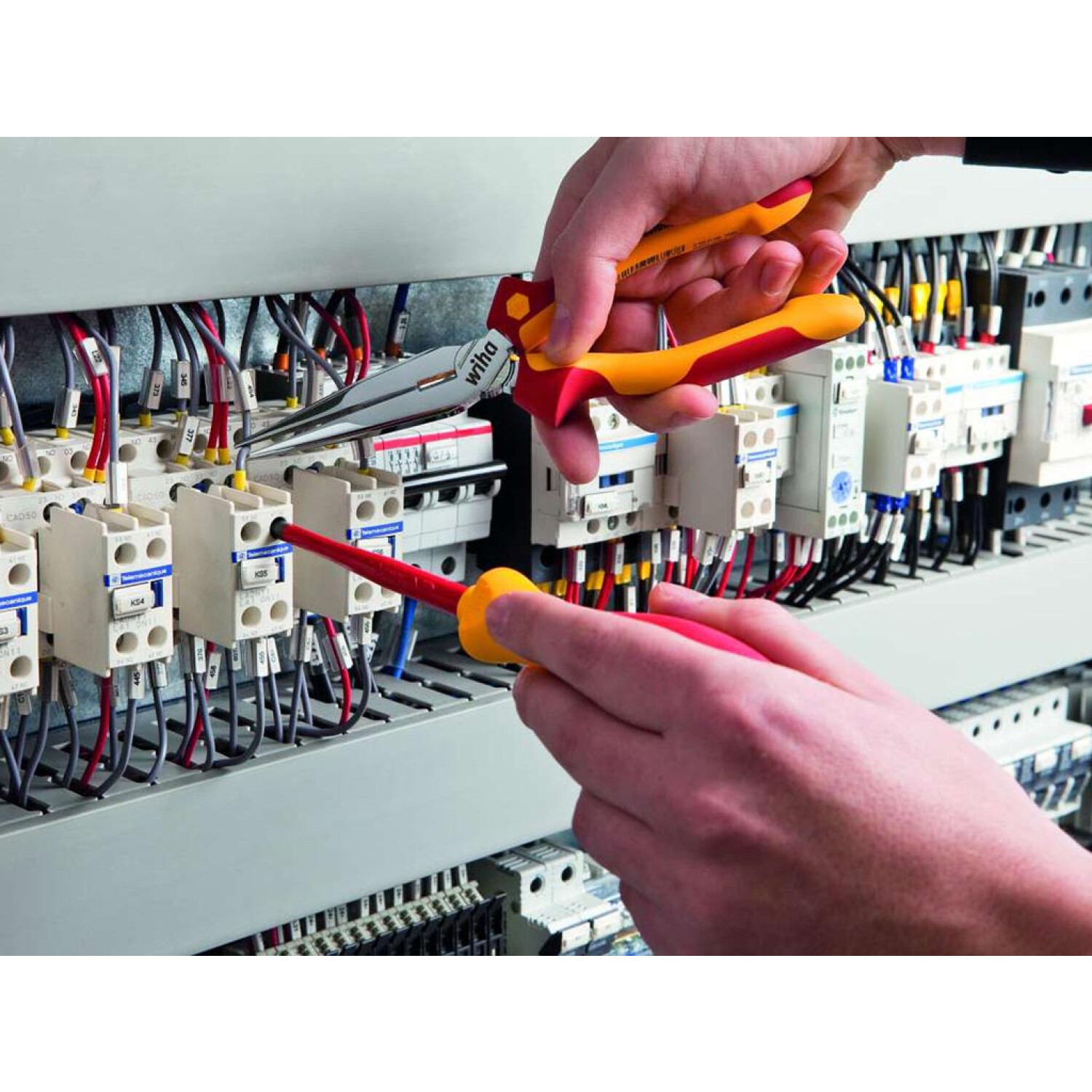 Osoba używająca czerwono-żółtych szczypiec i czerwonego śrubokręta na panelu elektrycznym.