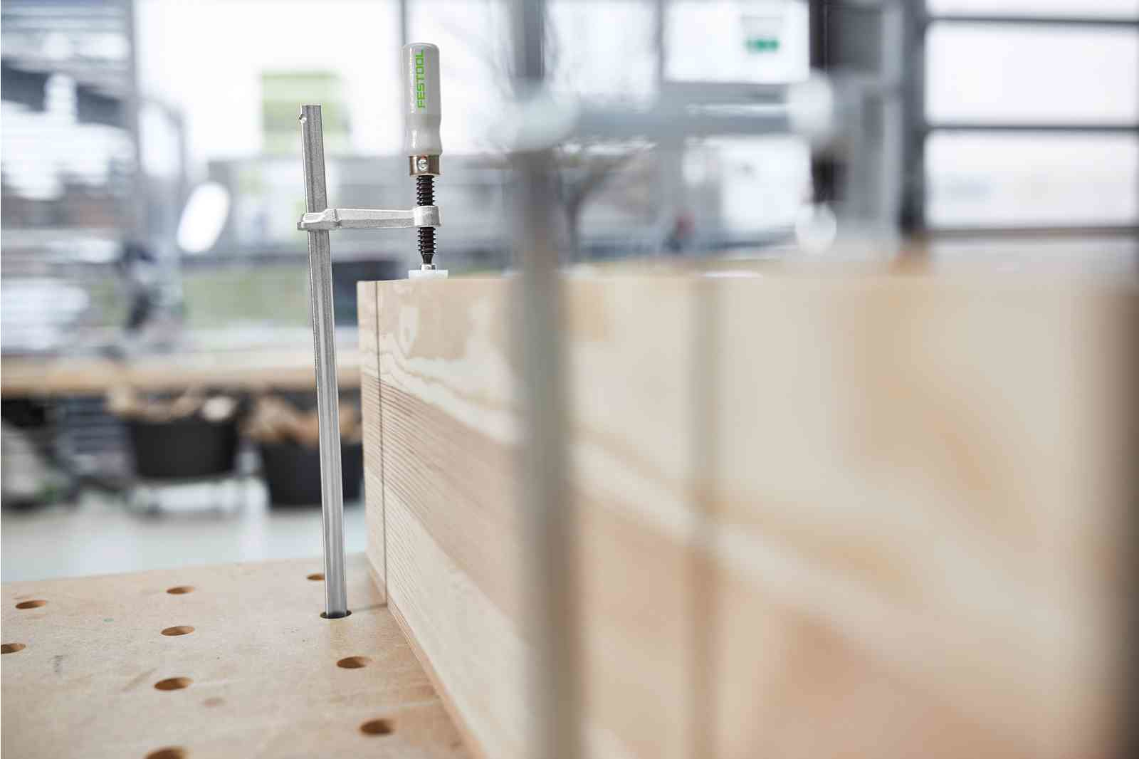 Zacisk mocuje kawałki drewna na stole warsztatowym. Rozmyte tło pokazuje okno i warsztat.