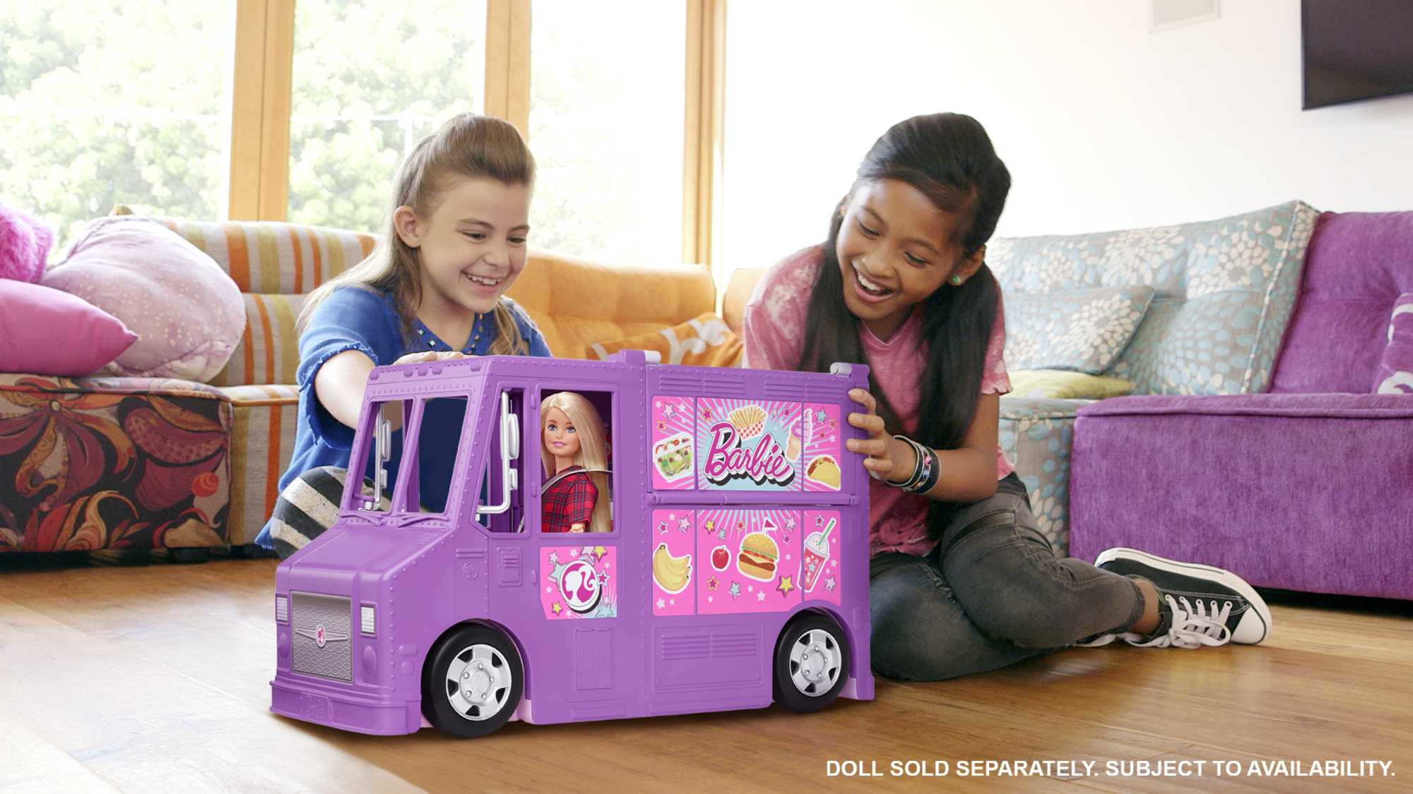 Zwei Mädchen lächeln und sitzen in der Nähe eines lila Barbie-Food-Trucks, mit einer Barbie-Puppe im Inneren.