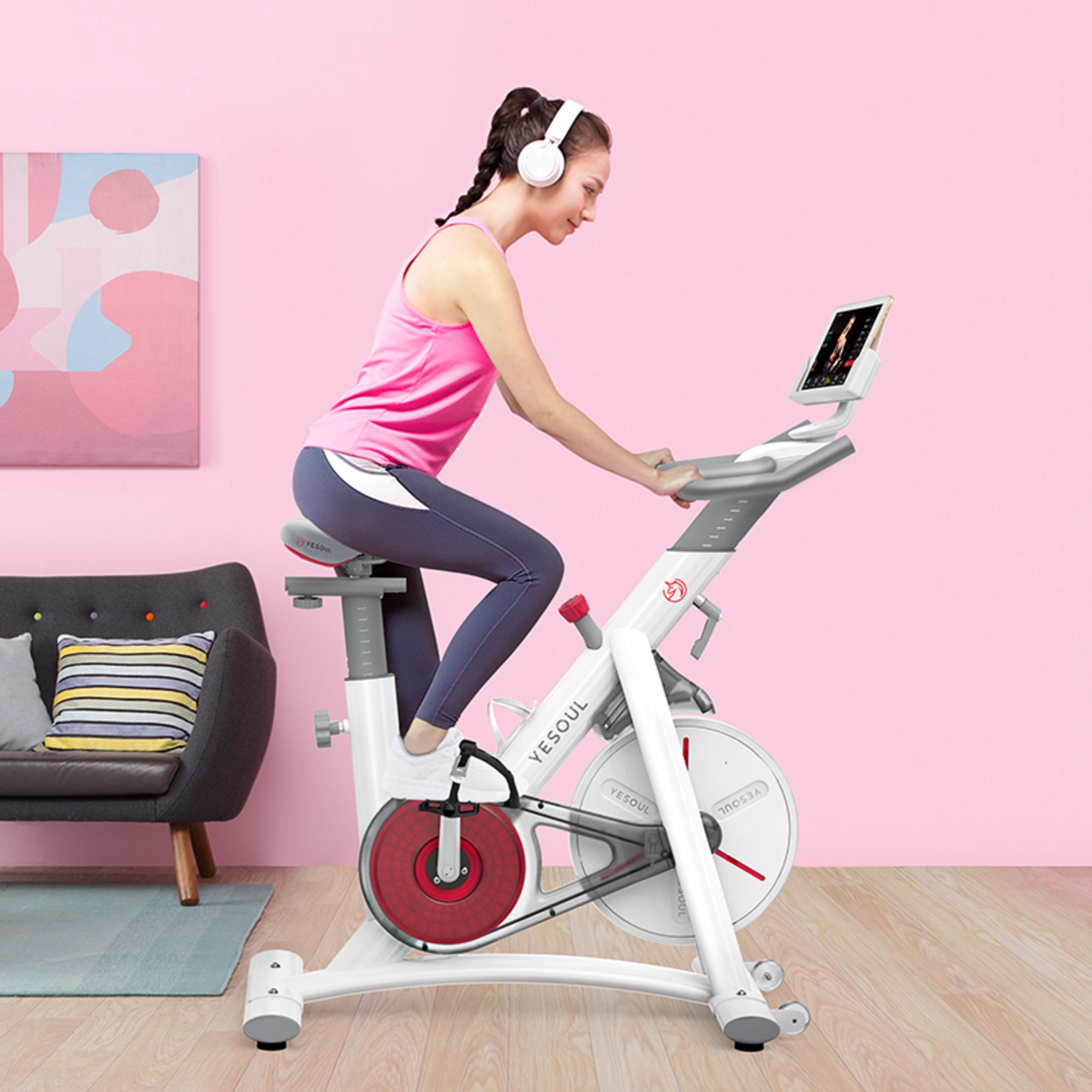 Donna su una cyclette bianca in una stanza rosa. Indossa cuffie e un top rosa.