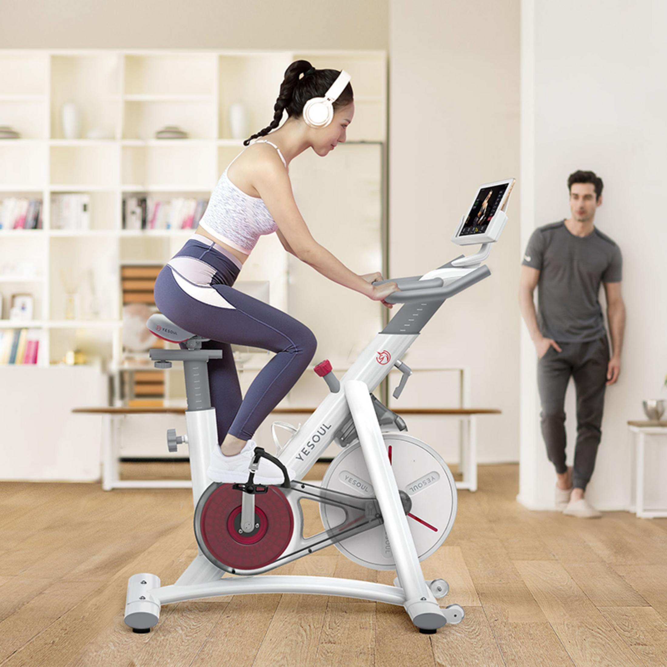 Donna su una cyclette, che si esercita. Un'altra persona cammina sullo sfondo. Bianco e rosso.