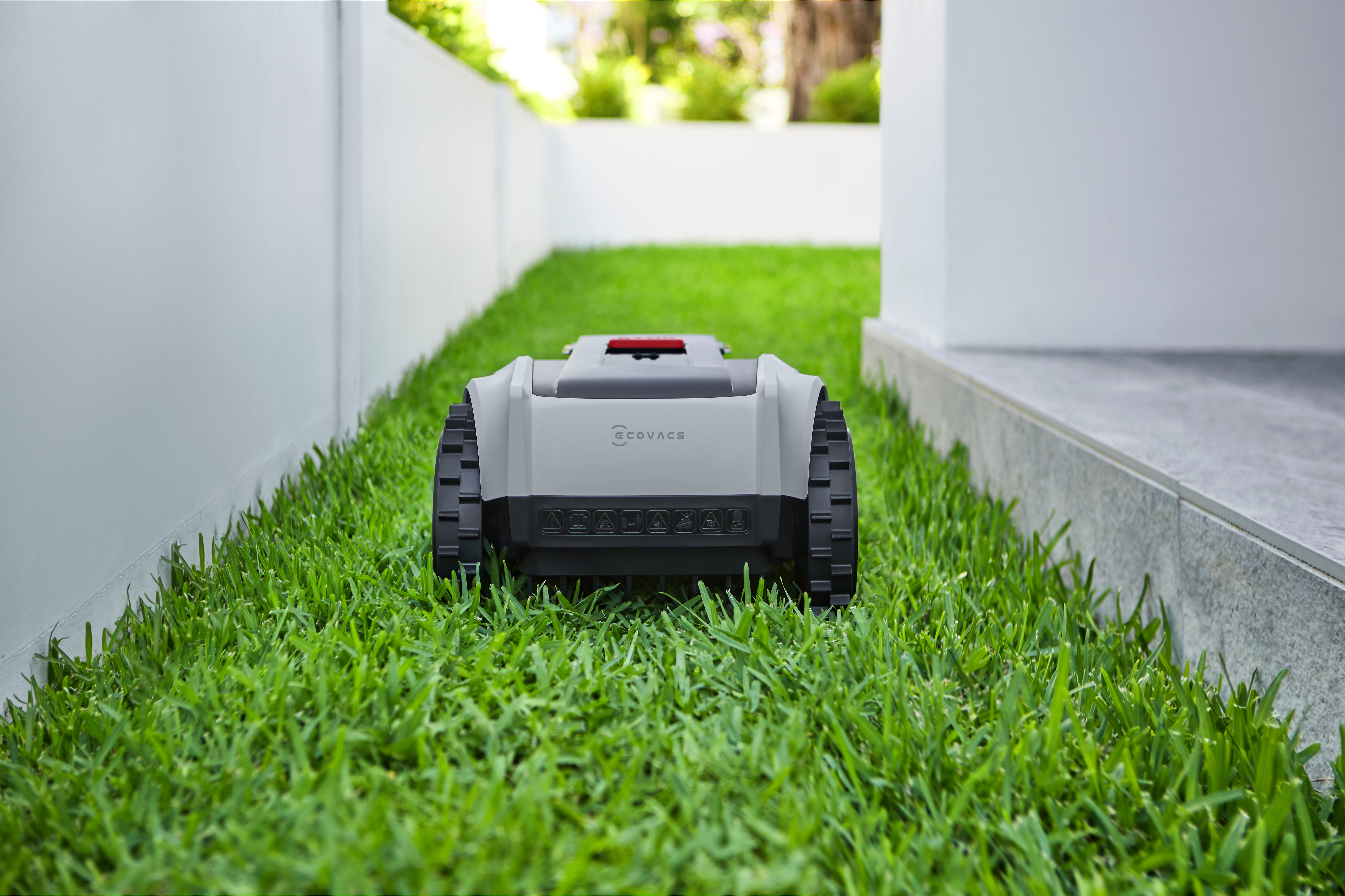 Ein Roboter-Rasenmäher auf grünem Gras. Der Mäher ist grau und schwarz. Weißer Zaun und Gebäude im Hintergrund.