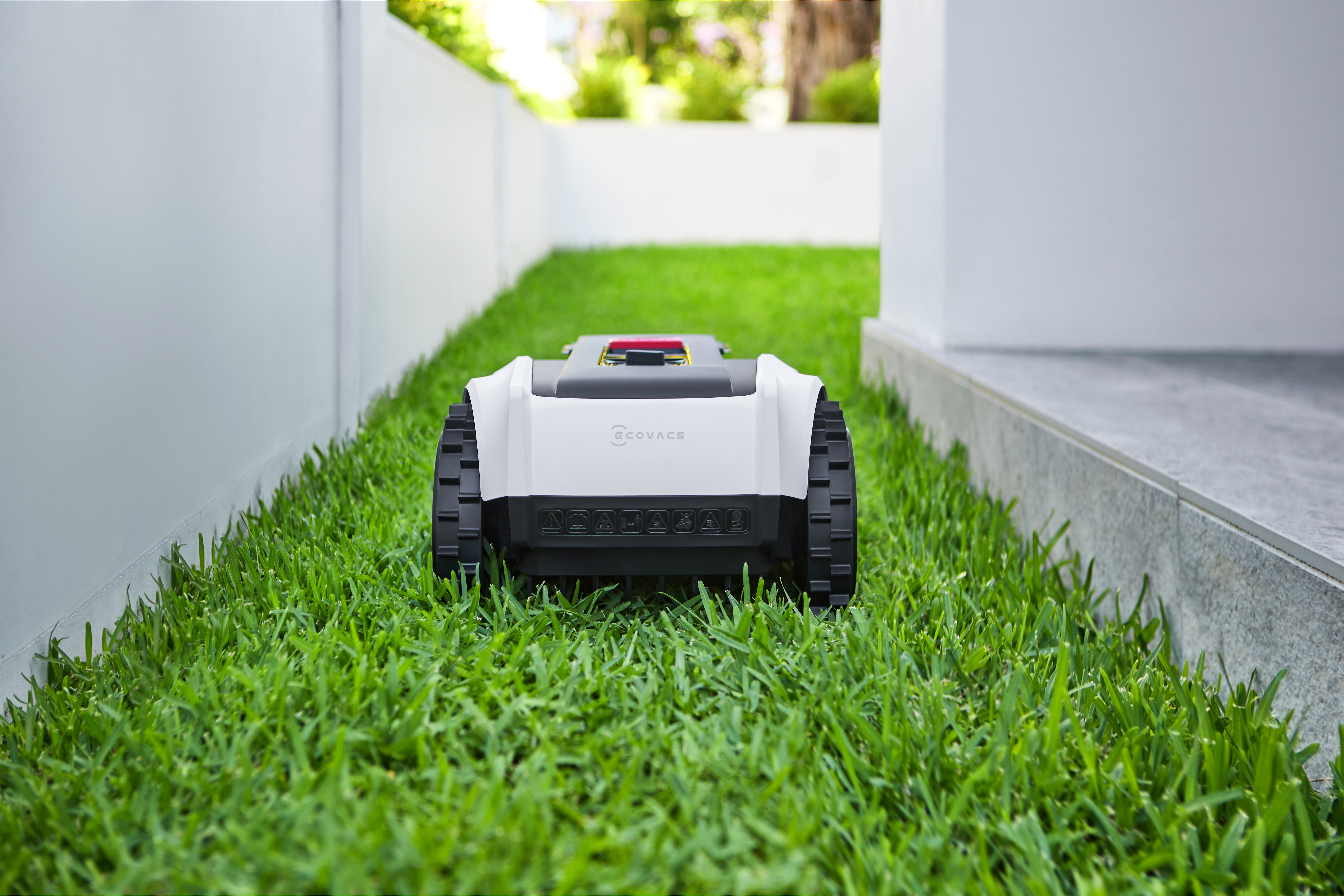 Biało-czarna kosiarka robot na zielonej trawie, w pobliżu białej ściany i chodnika.