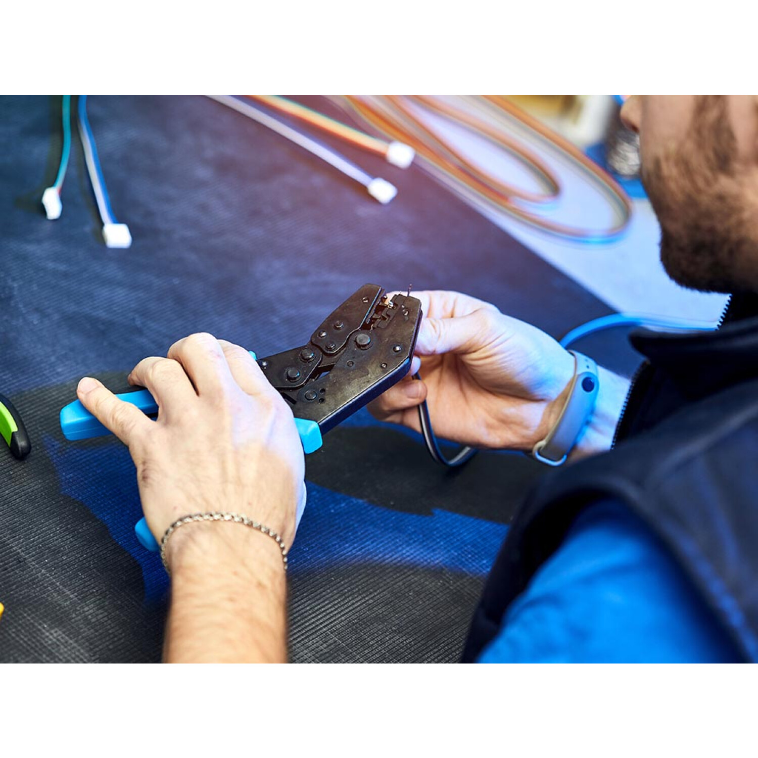 Osoba używająca zaciskarki z niebieskim uchwytem na przewodach. Widoczna opaska na nadgarstek.