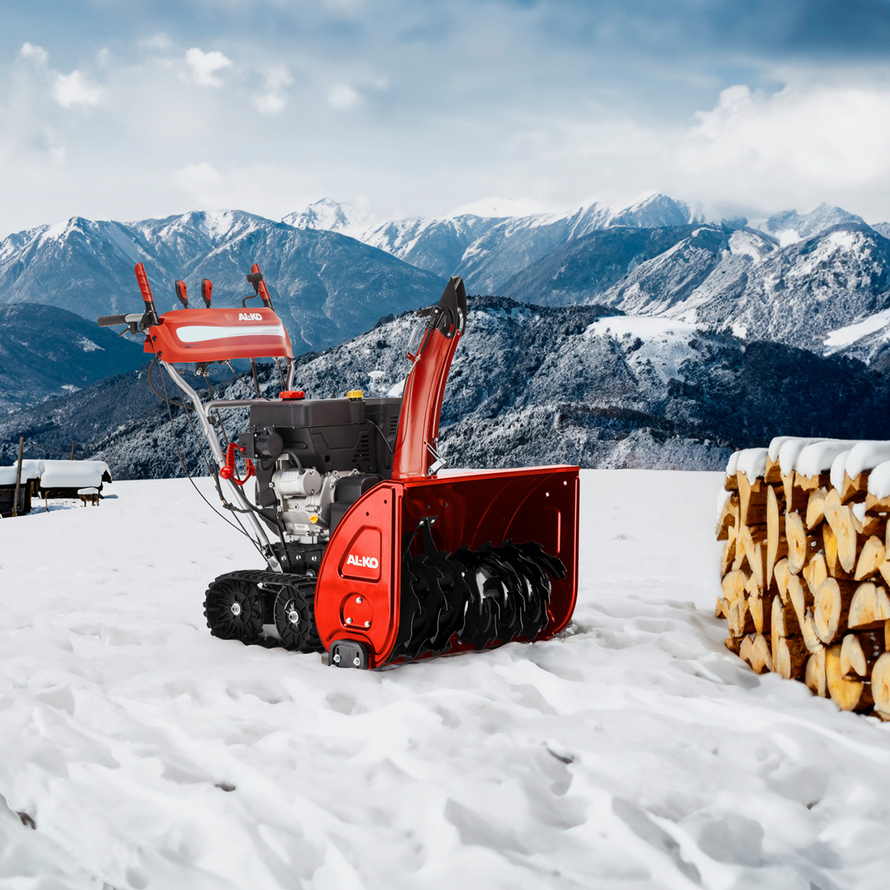 Roter Schneefräse im Schnee mit Bergkulisse und einem Holzhaufen.