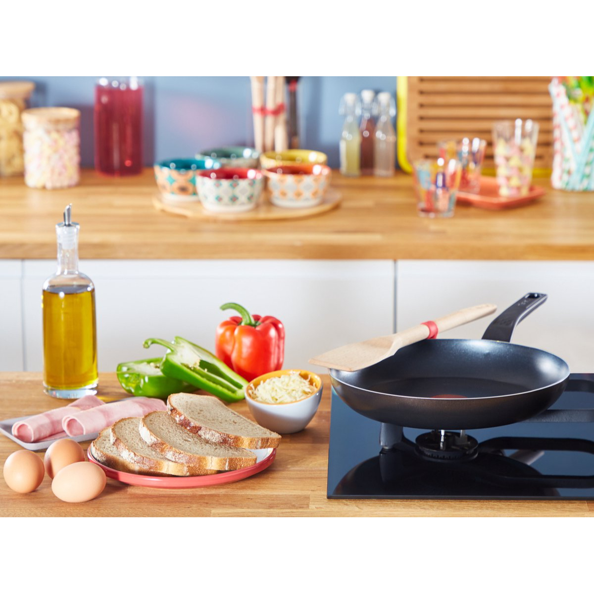 Patelnia Tefal na czarnej płycie grzewczej, otoczona produktami spożywczymi, w tym jajkami i warzywami.