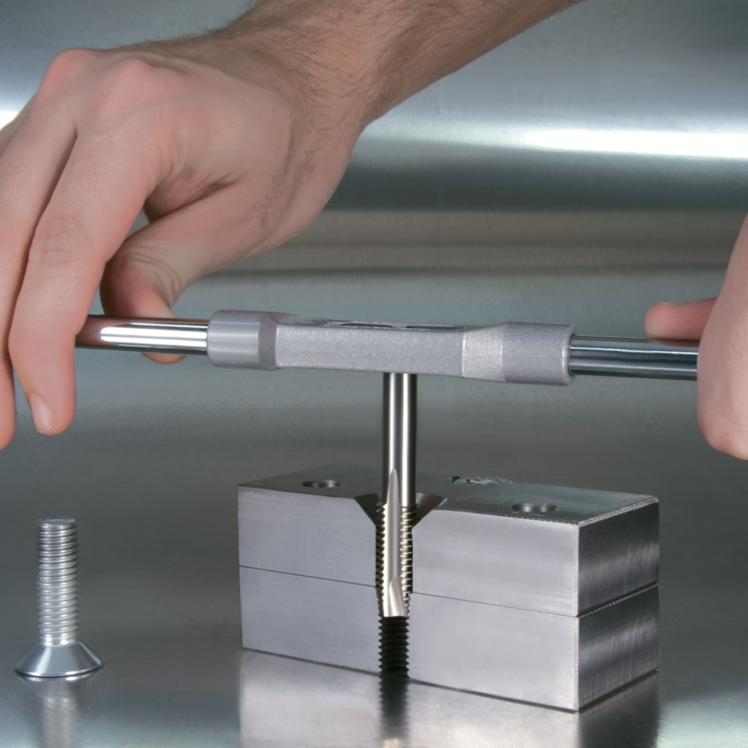Hände verwenden einen Gewindebohrer, um einen Metallblock zu bearbeiten. Eine Schraube ist in der Nähe.