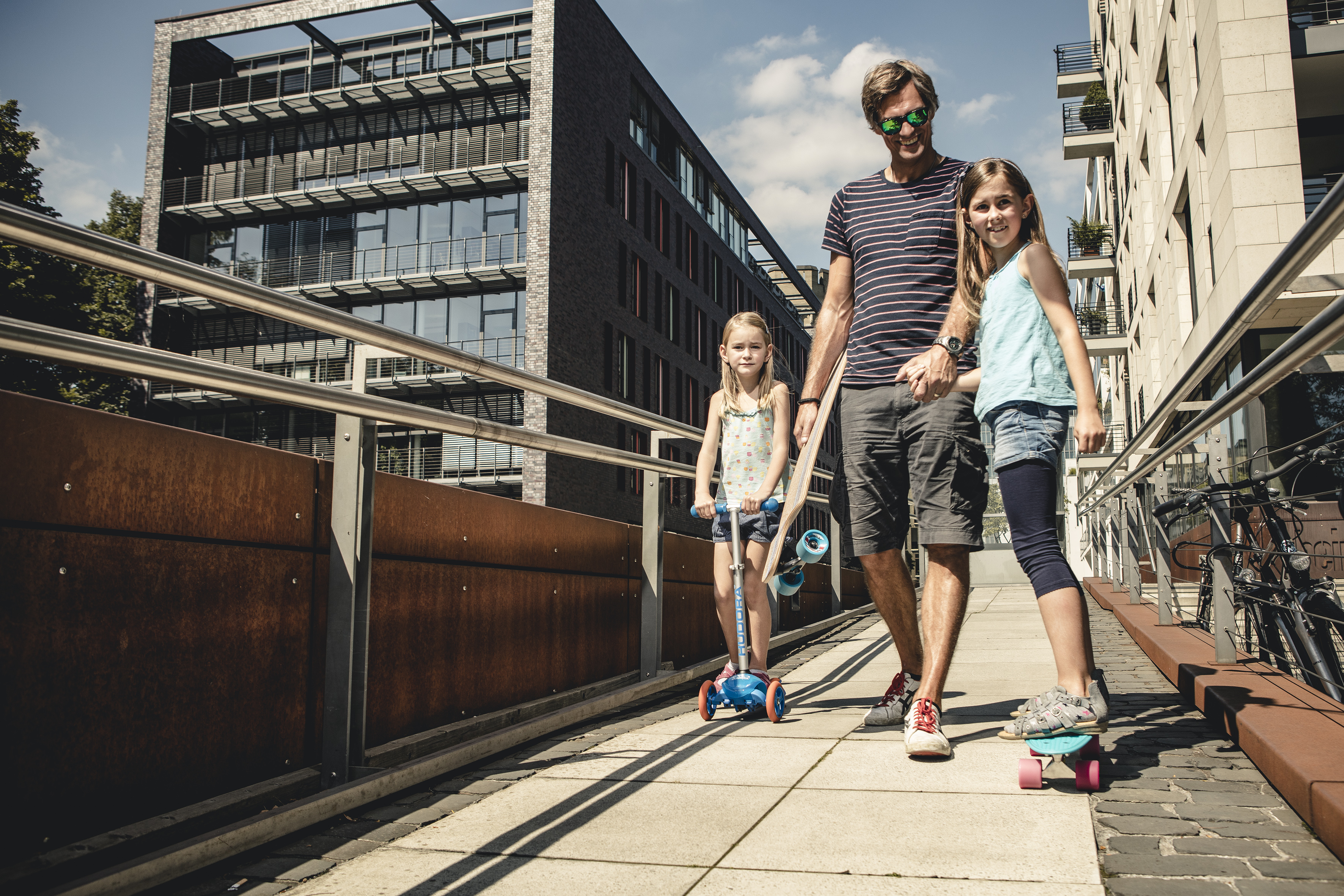 Un uomo e due ragazze camminano su un ponte con scooter e skateboard.