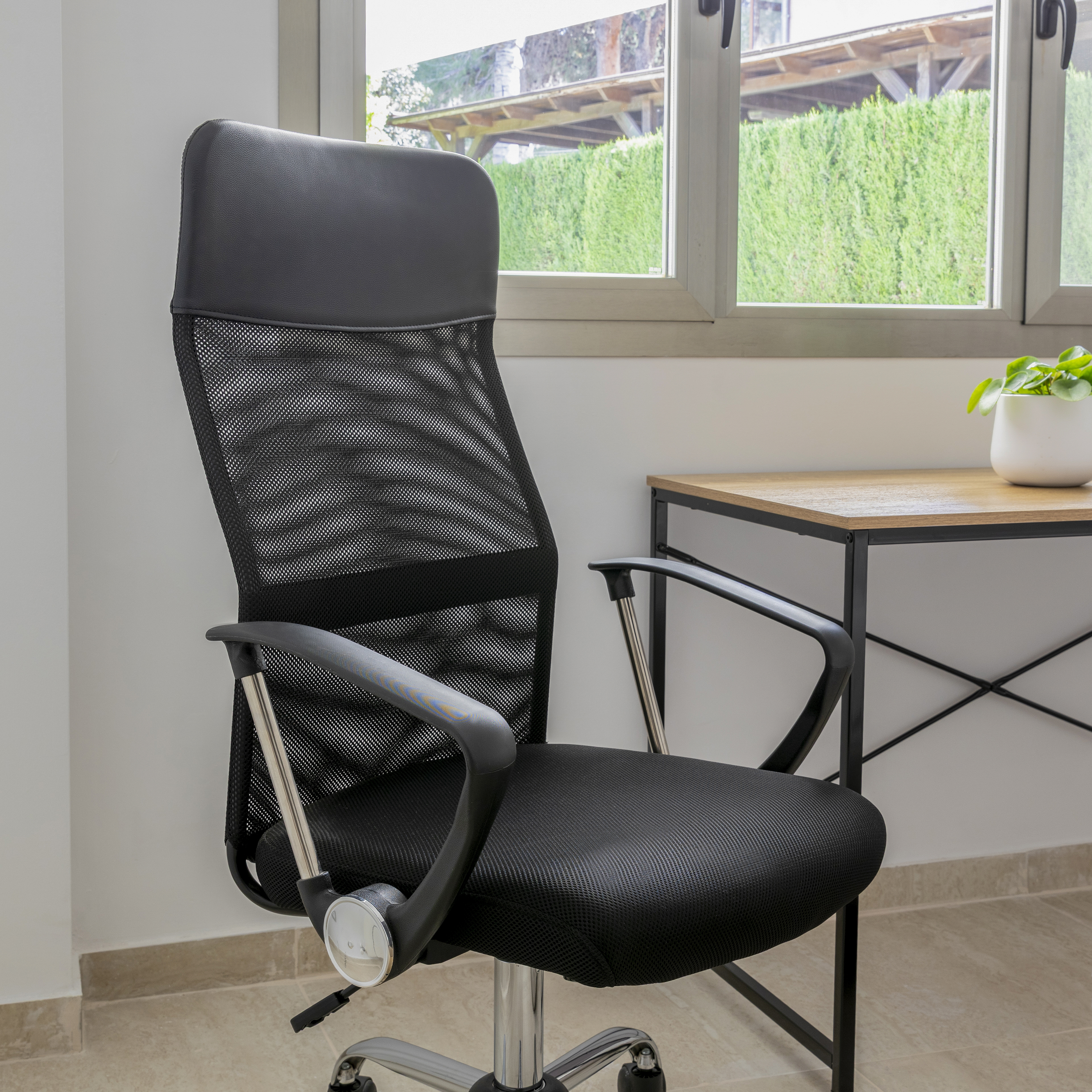 Silla de oficina negra de malla, marco plateado, cerca de una ventana, escritorio de madera y planta verde.