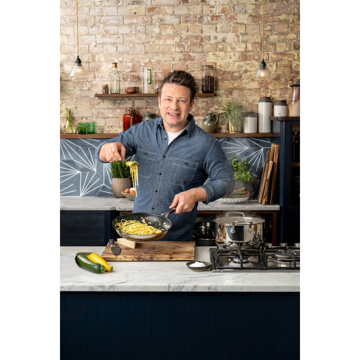 Jamie Oliver kookt pasta in een pan, met ingrediënten en een bakstenen muur op de achtergrond.