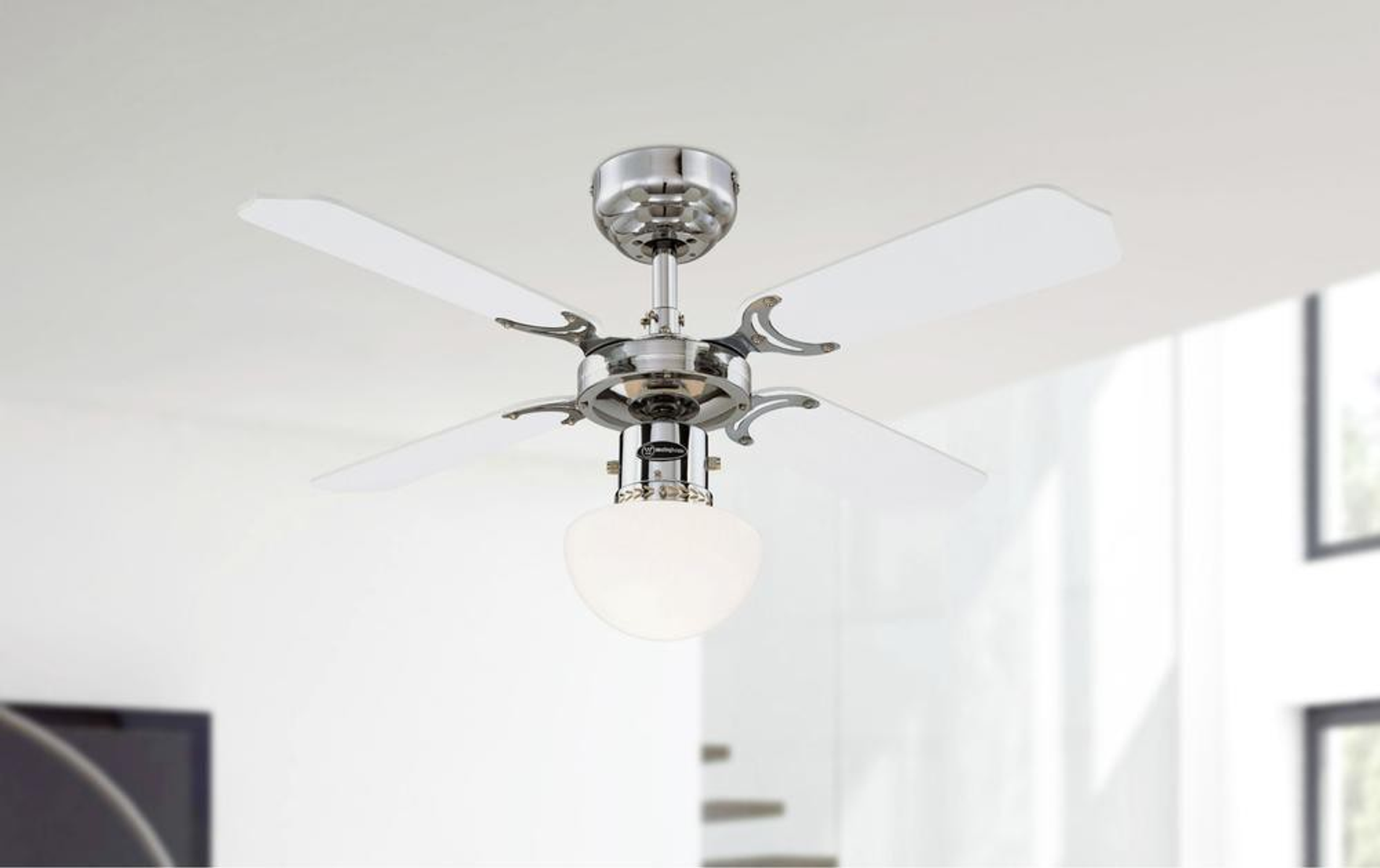 Ein Deckenventilator mit vier weißen Flügeln und einer zentralen Leuchte. Die Basis ist verchromt.